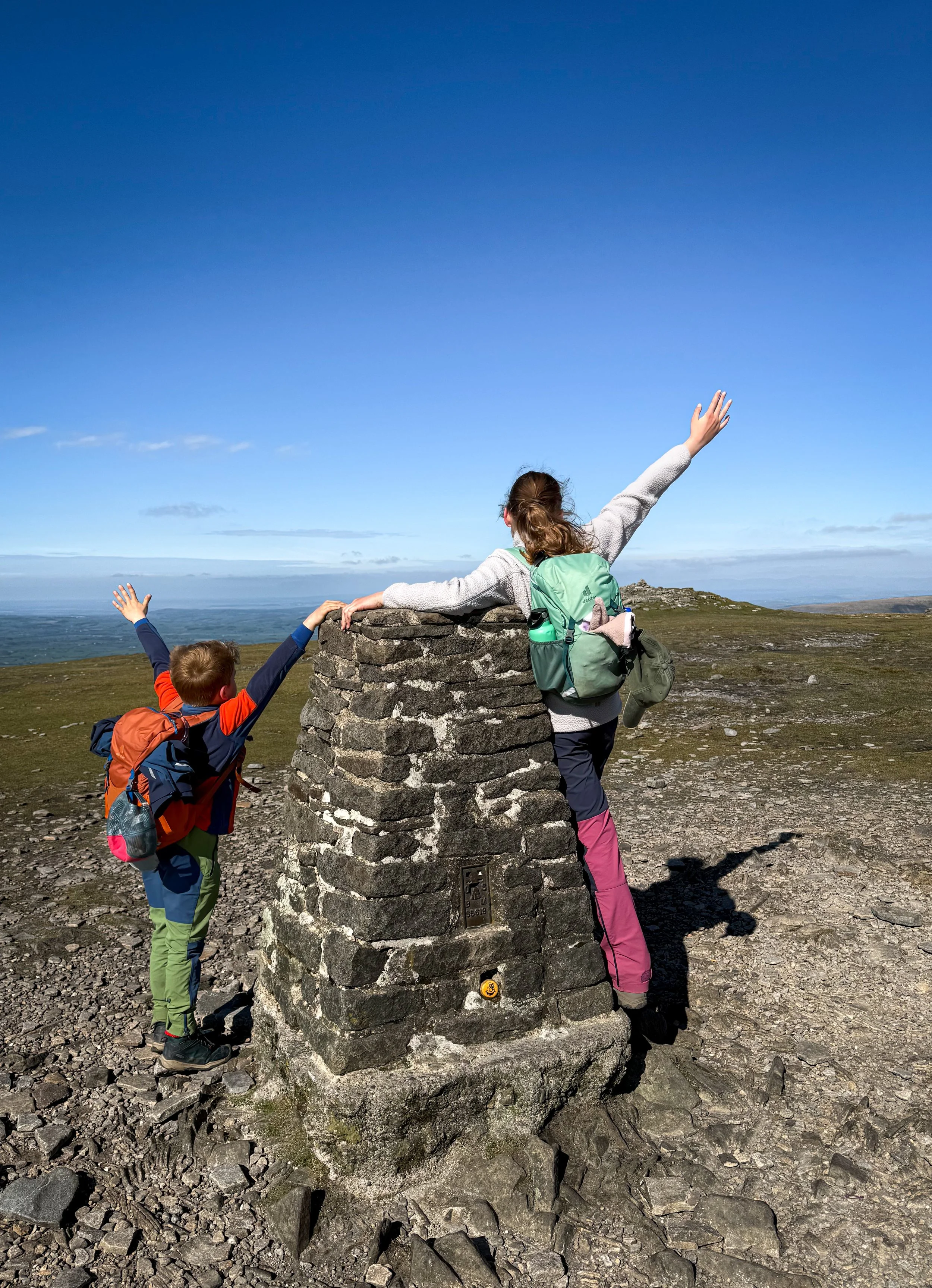 Children standing at a trig point with arms in the air