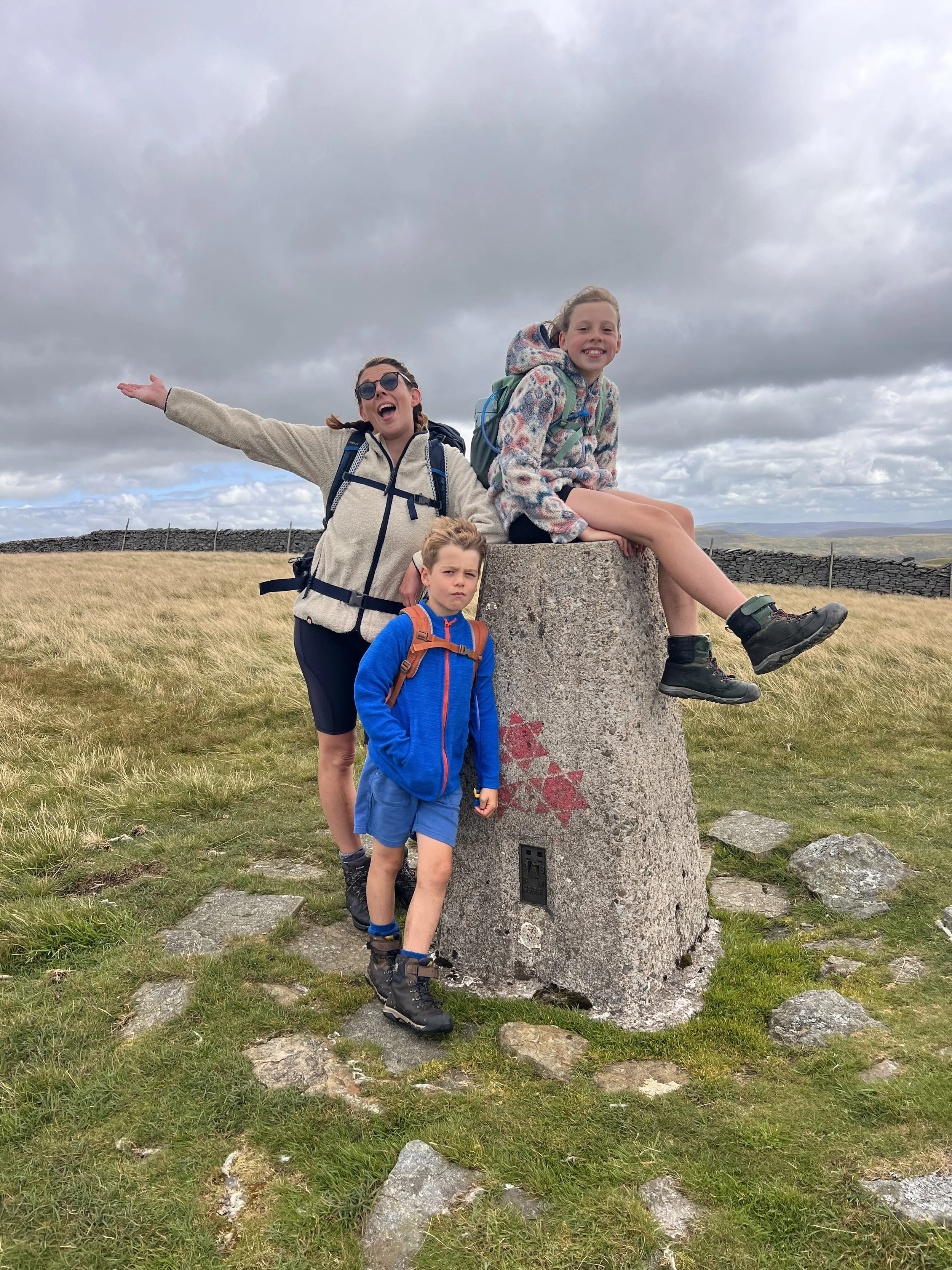 An adult and two children sitting on and standing next to a trig point decorated with red stars and with a dry stone wall behind