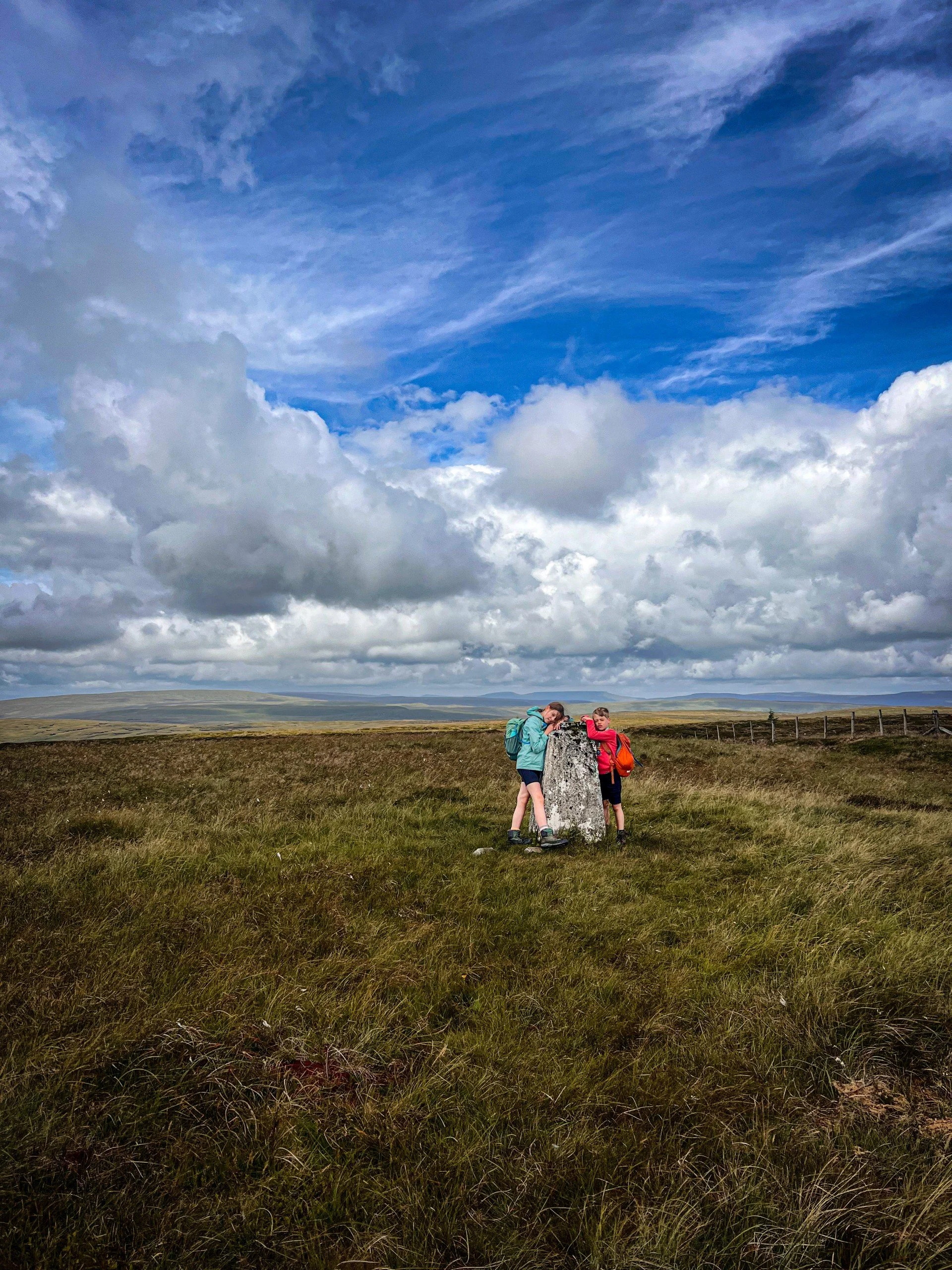 Children on top of a moor with a trig point under blue skies in Summer