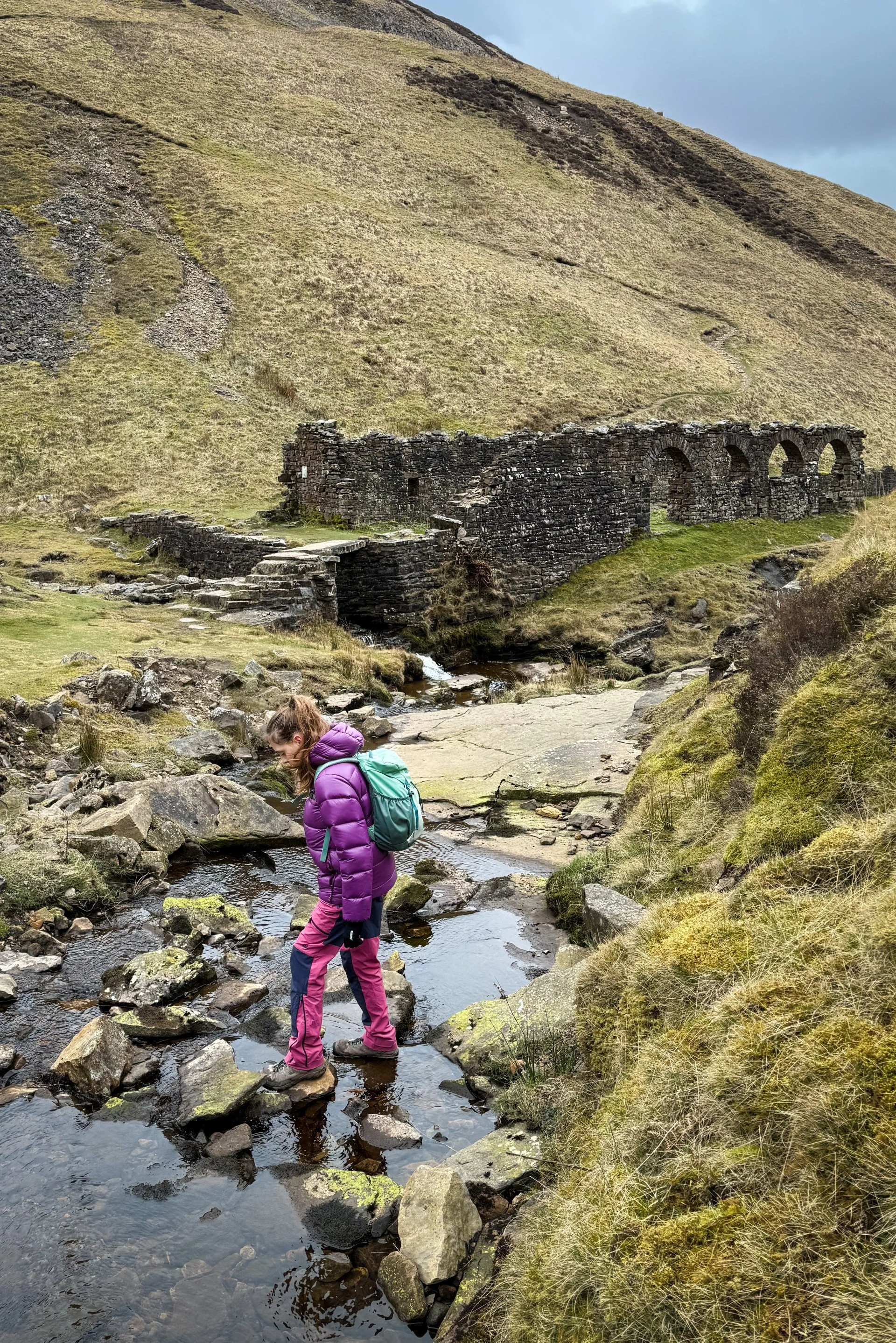 Child walking across a stream wit ruined mine buildings behind wearing a purple coat and pink trousers