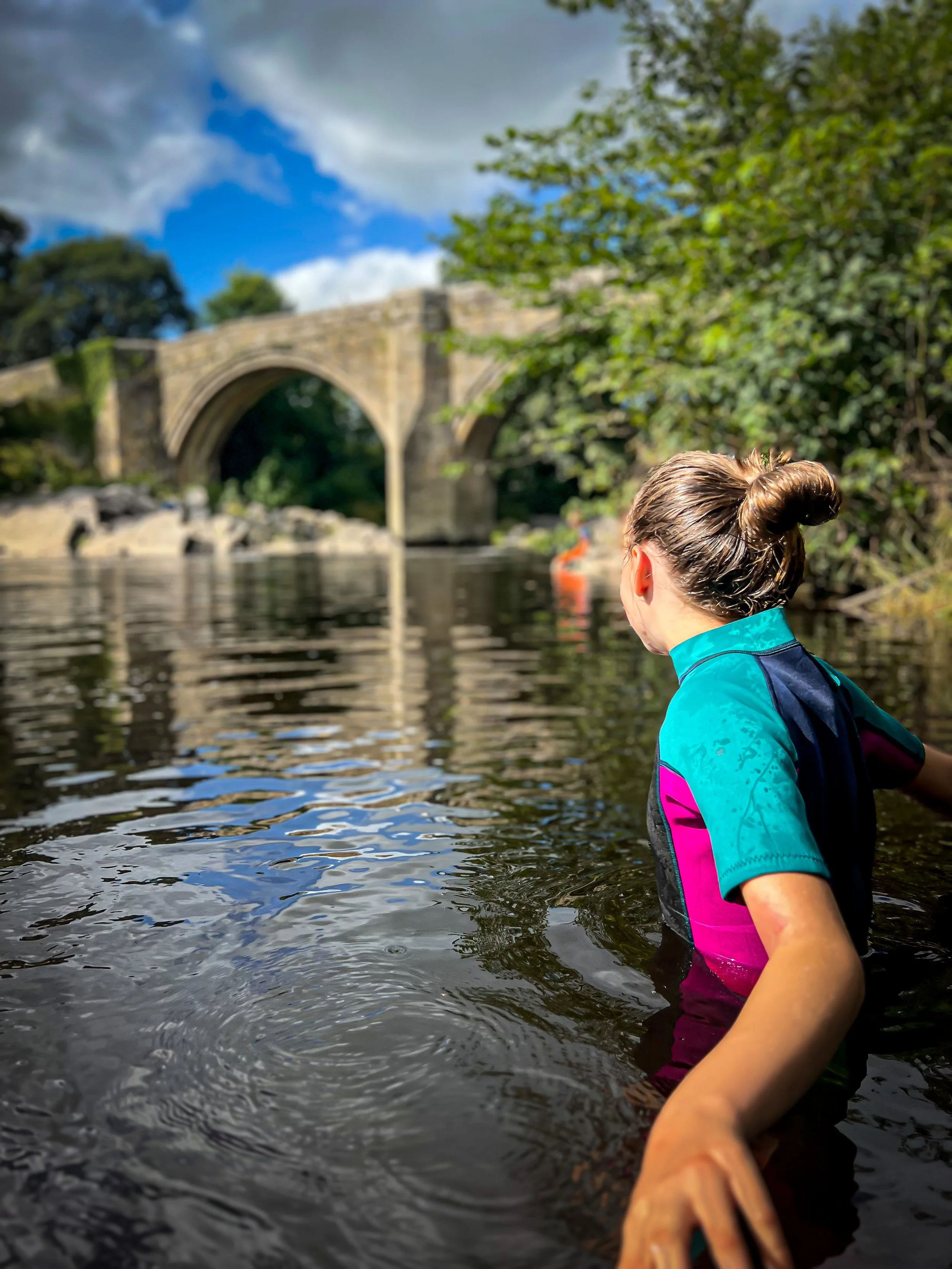 A child in the river at an old bridge in summer with green trees