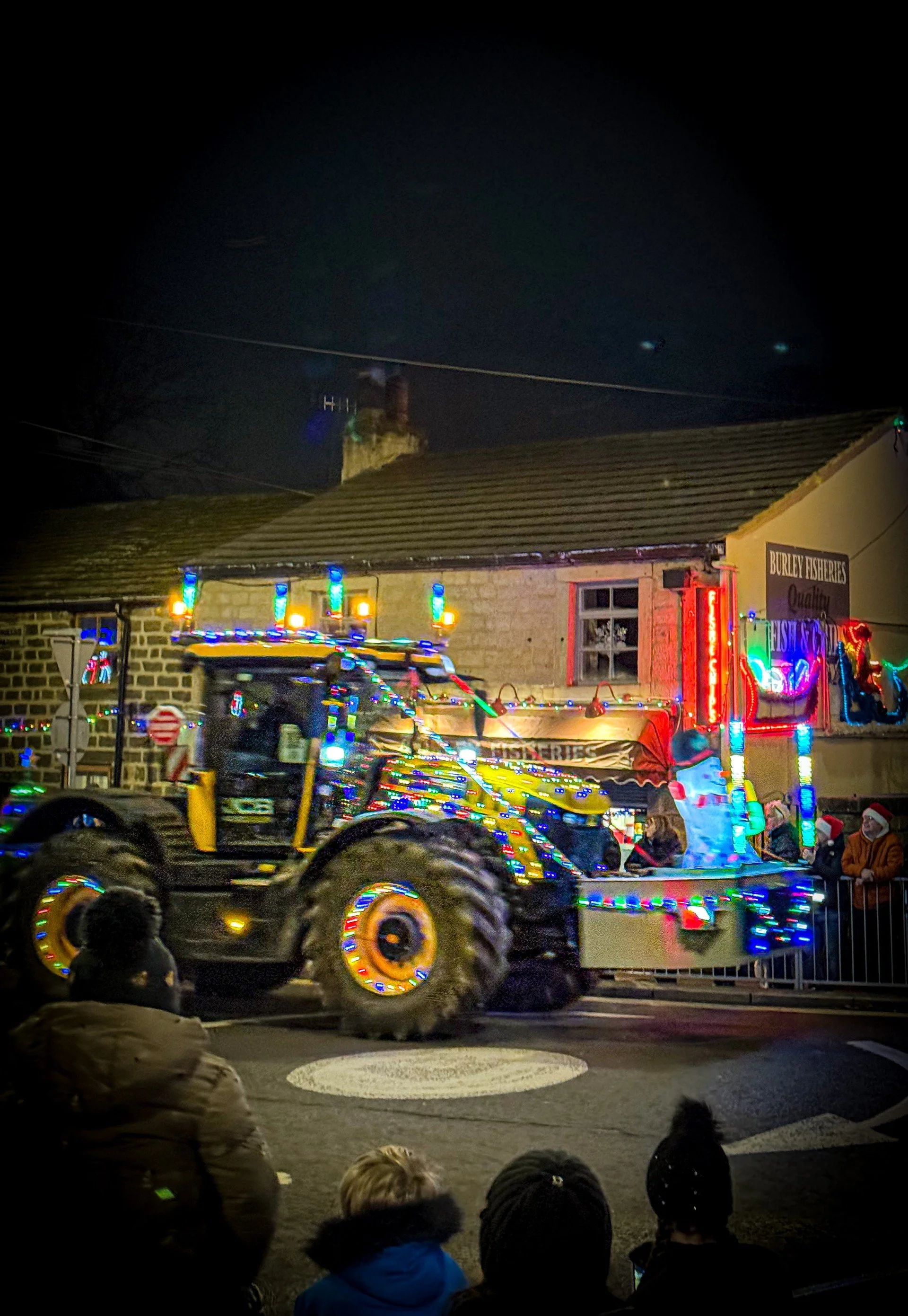 A tractor driving through a village covered in Christmas lights
