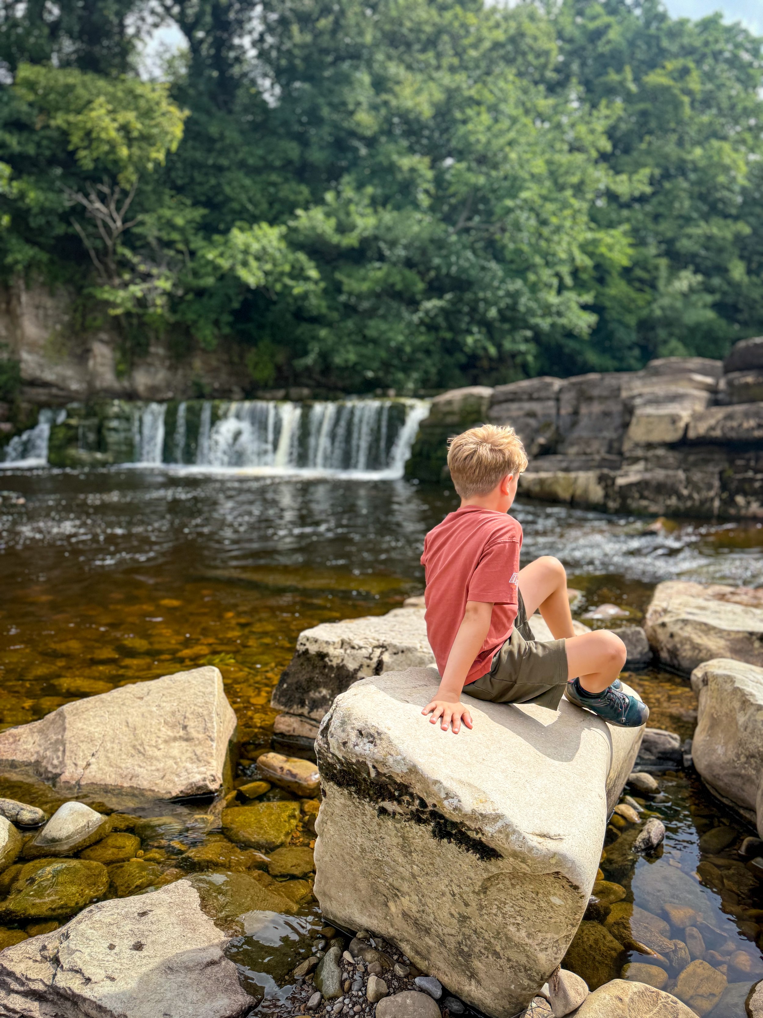 Child sat on a rock next to a river with a large waterfall behind