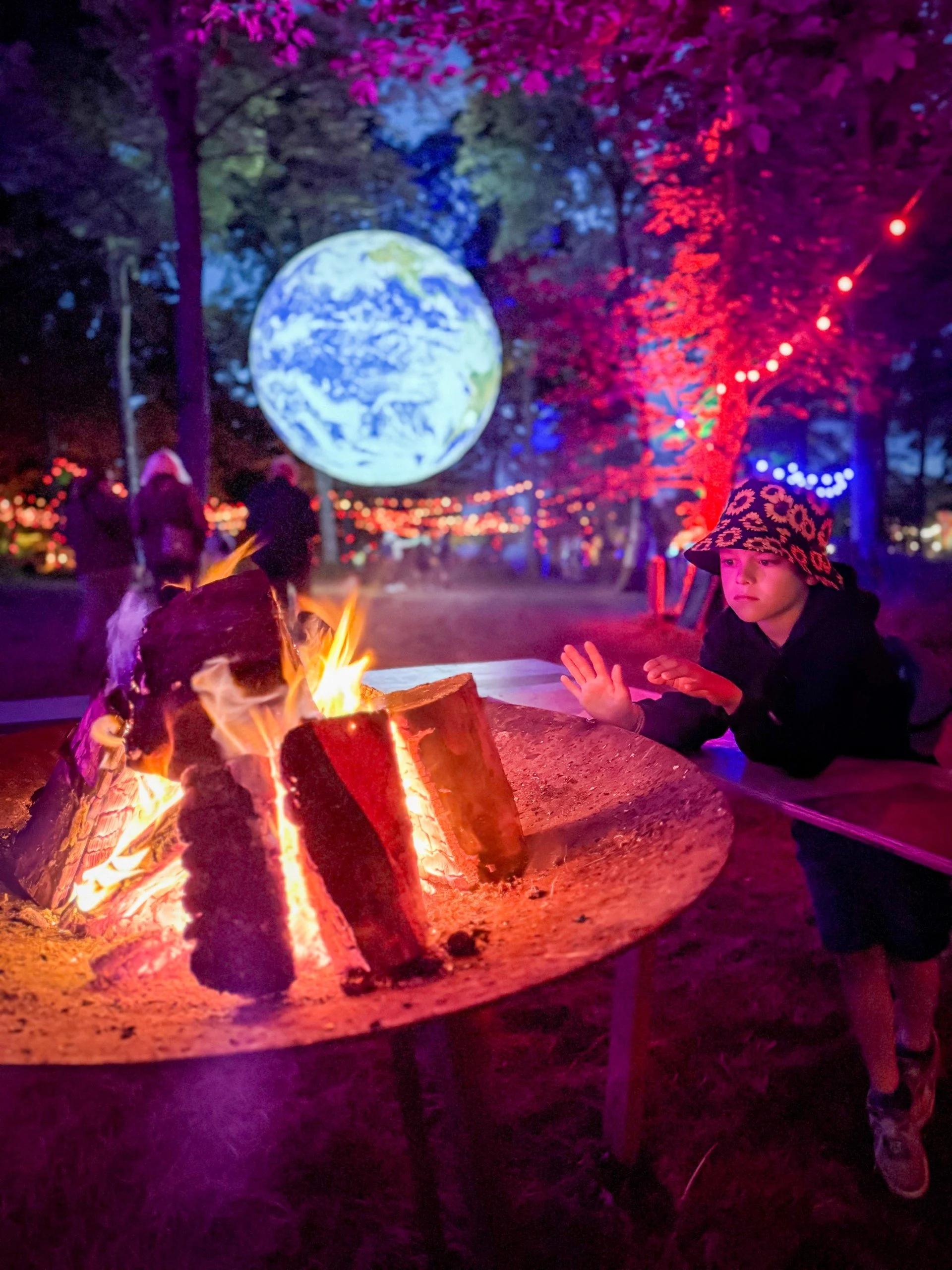Child sitting by a fire pit with an illuminated earth sculpture behind in woodland thats lit up with red and purple