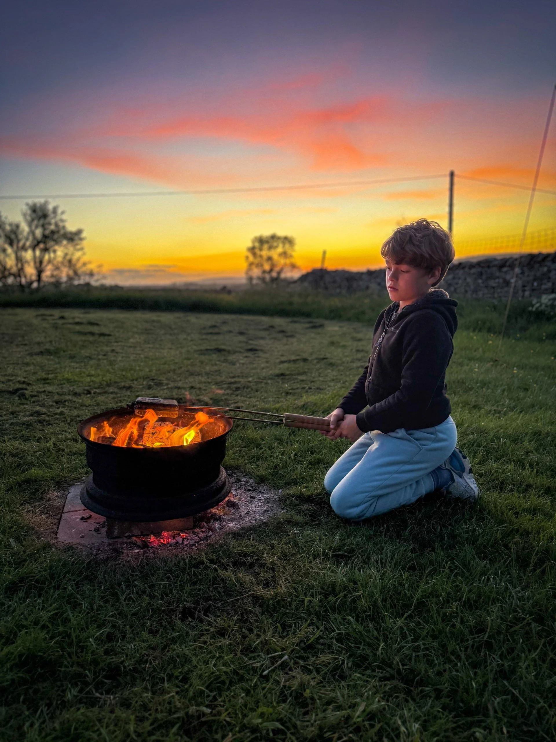 Child sitting next to  campfire toasting something with sunset skies behind and grass beyond
