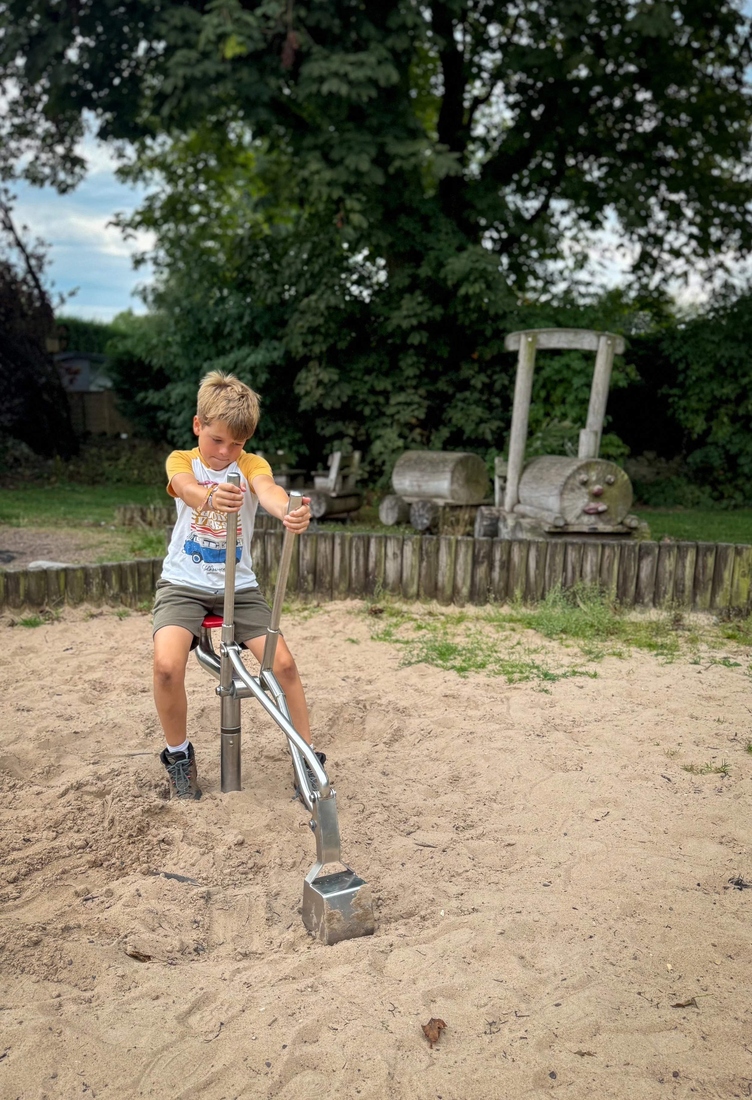 Child sitting on a metal play digger in a sand pit with a wooden train in the background