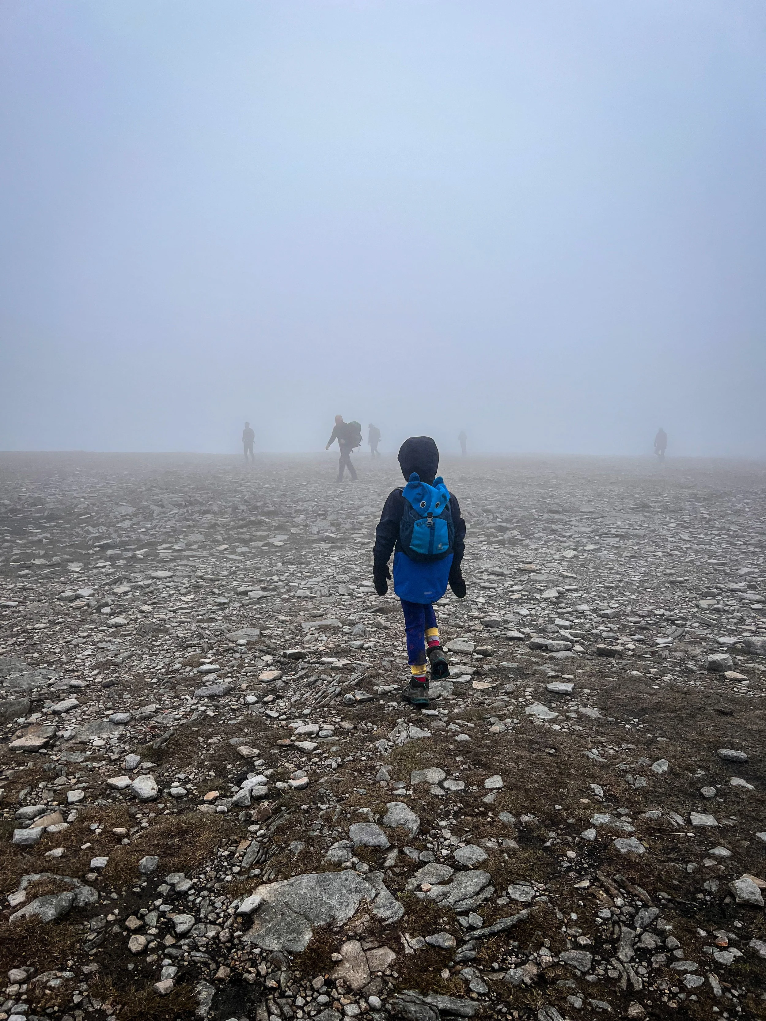 Child walking in cloud on top of a flat rocky plateau with shadows of people ahead