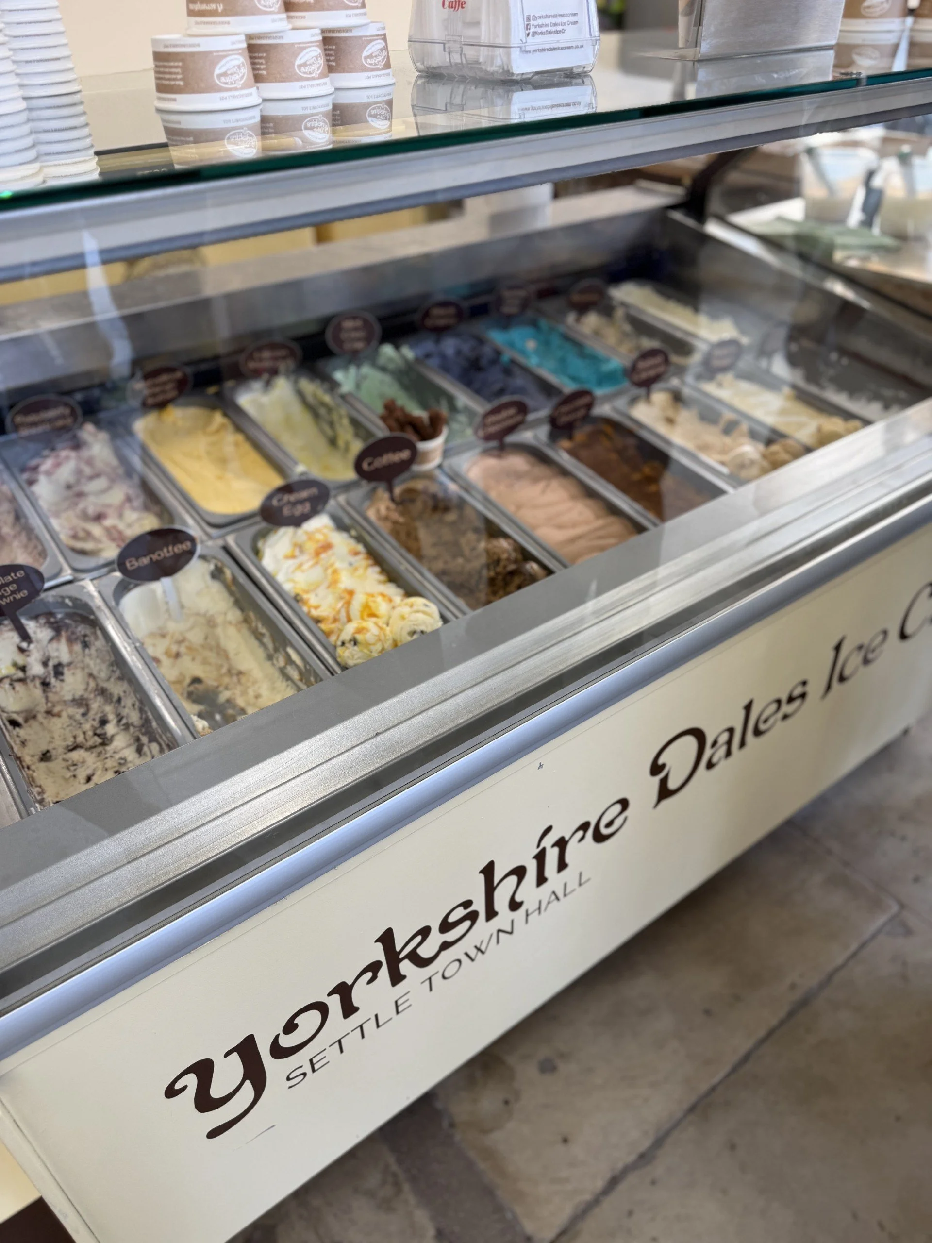 lots of tubs filled with lots of different kinds of ice cream all different colours inside a freezer with tubs piled on top
