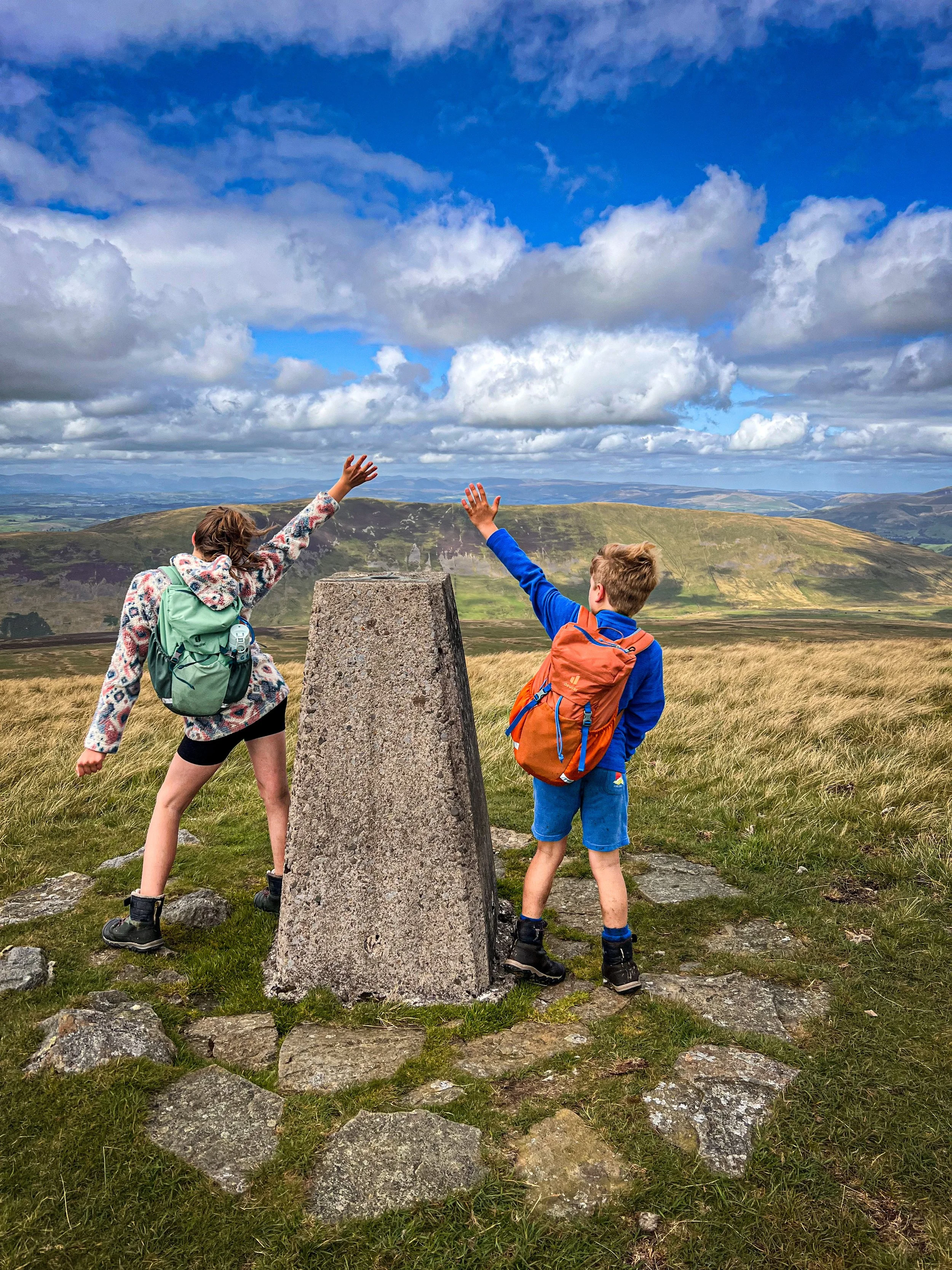 Children standing next to a trig point looking out at the view beyond to mountains under blue skies