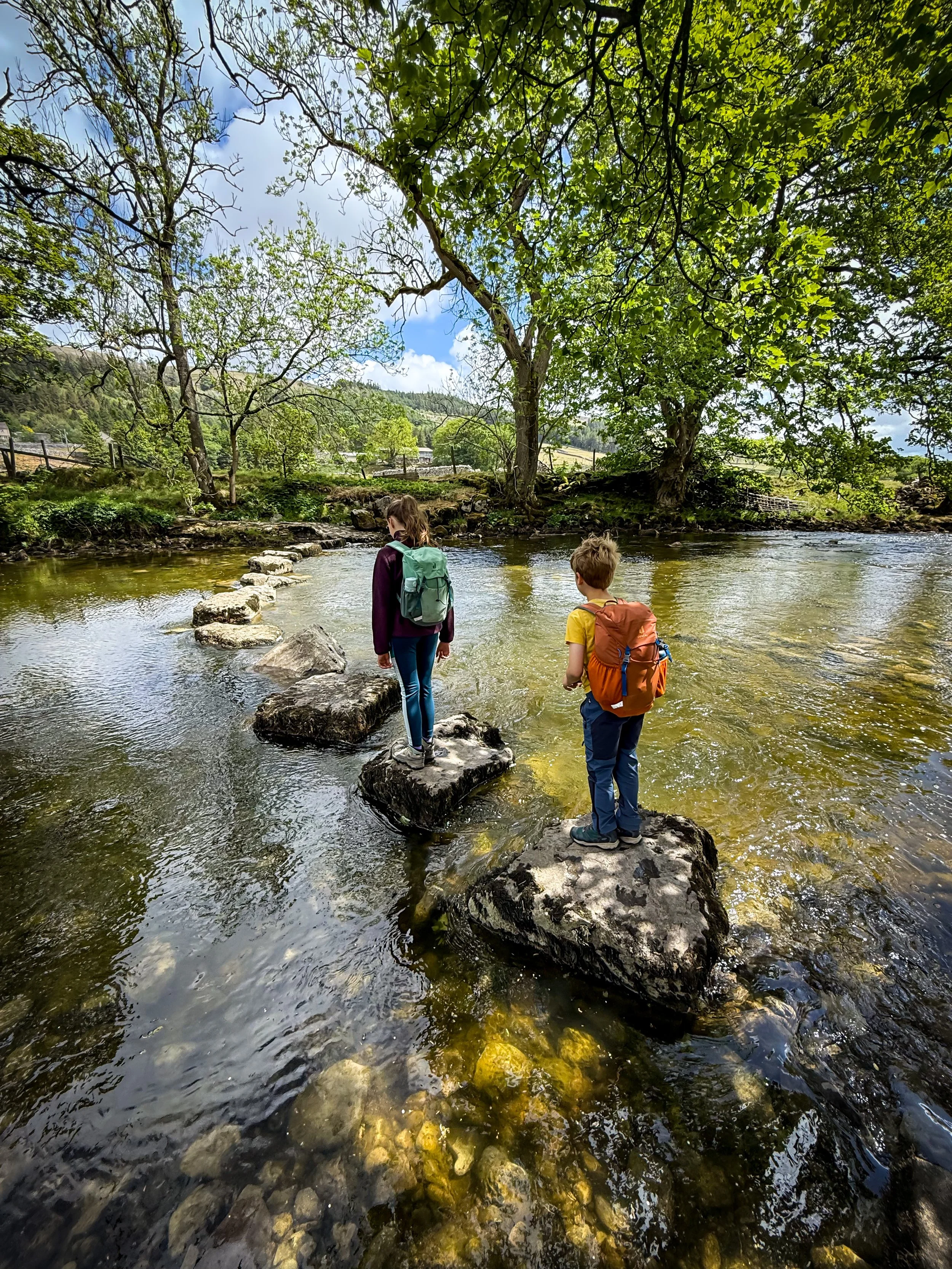 Children walking across stone stepping stones under trees with river