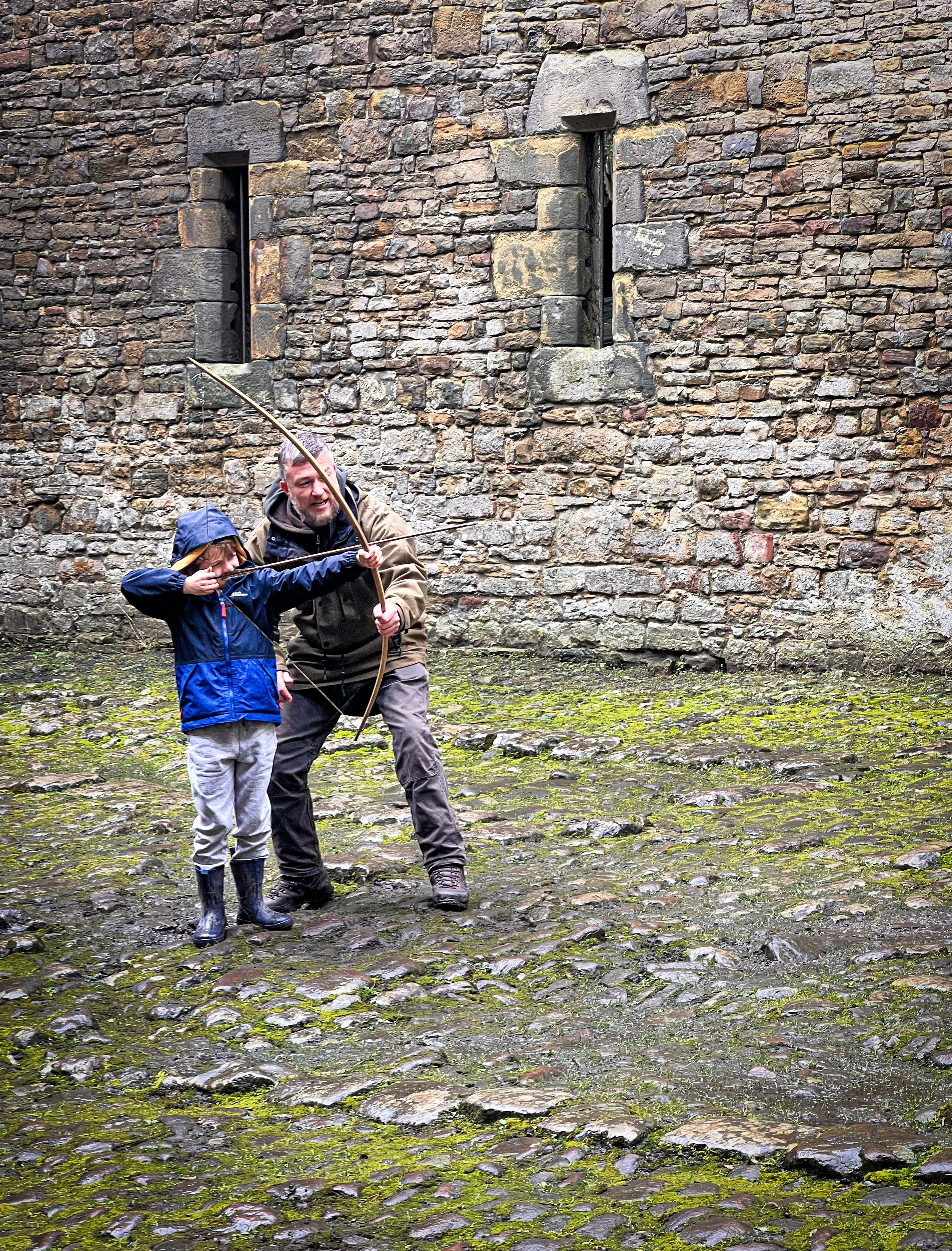 Child with bow and arrow being guided by adult to fire with castle behind