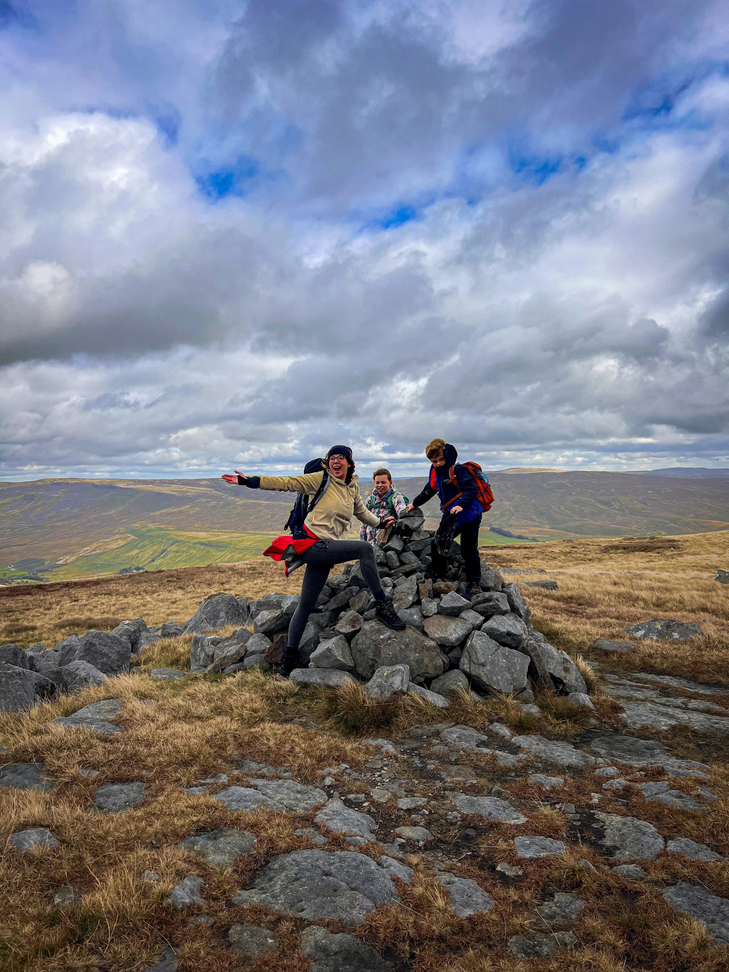 An adult and two children on a summit cairn with mountains behind and blue cloudy skies above