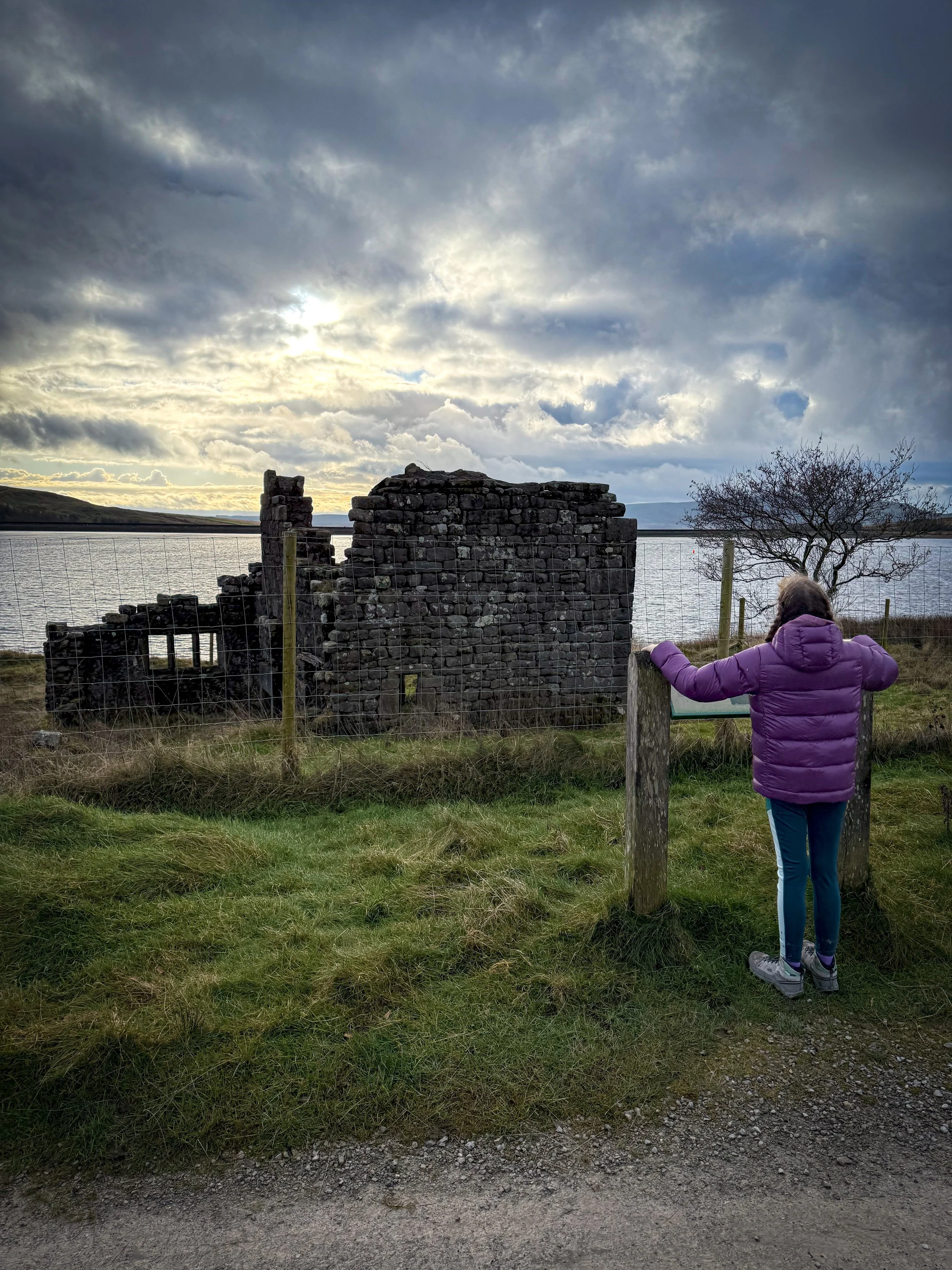Child standing at the ruins of one of the old village buildings with reservoir behind