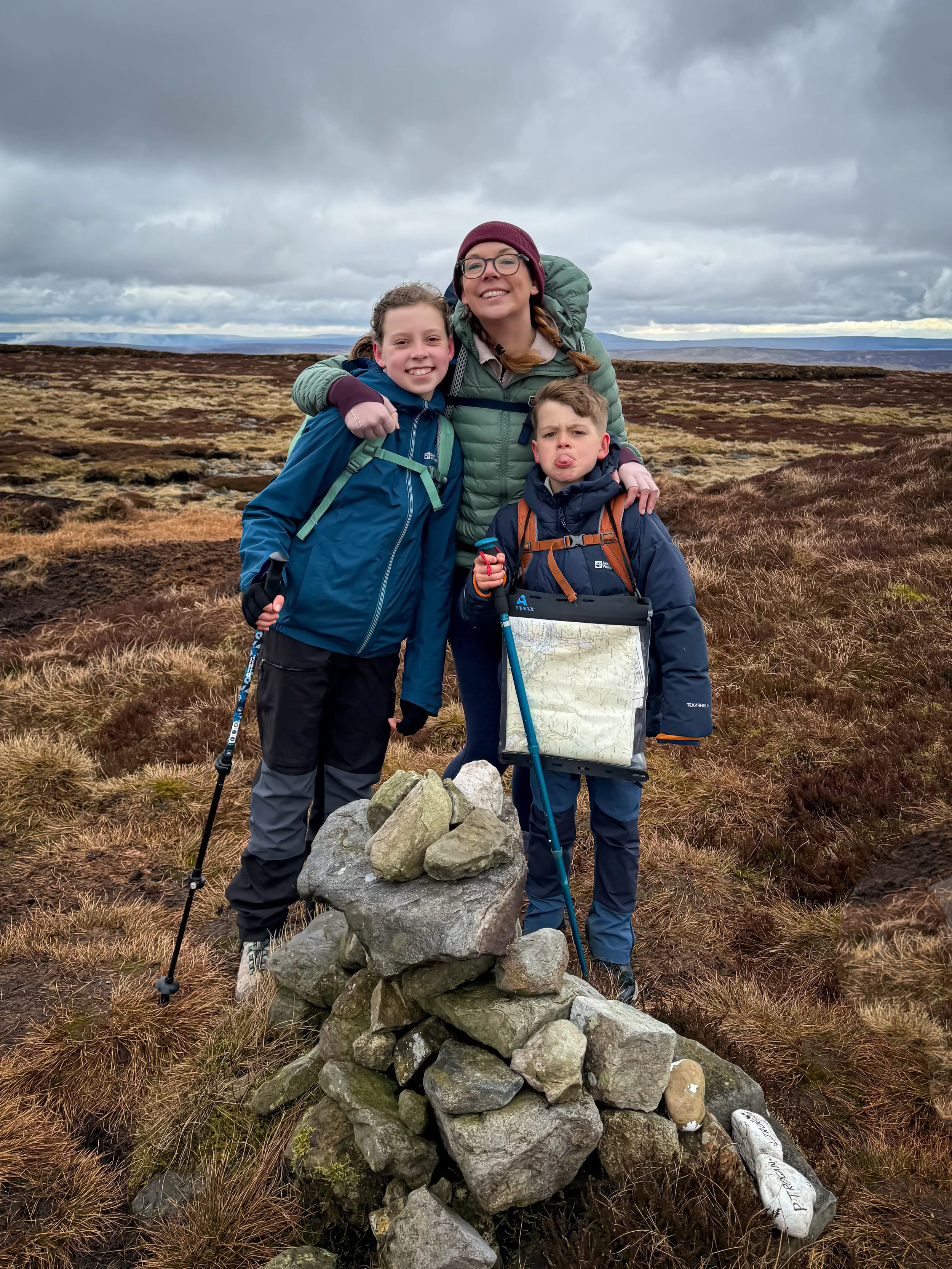 An adult and two children standing at a summit cairn with a map and walking poles