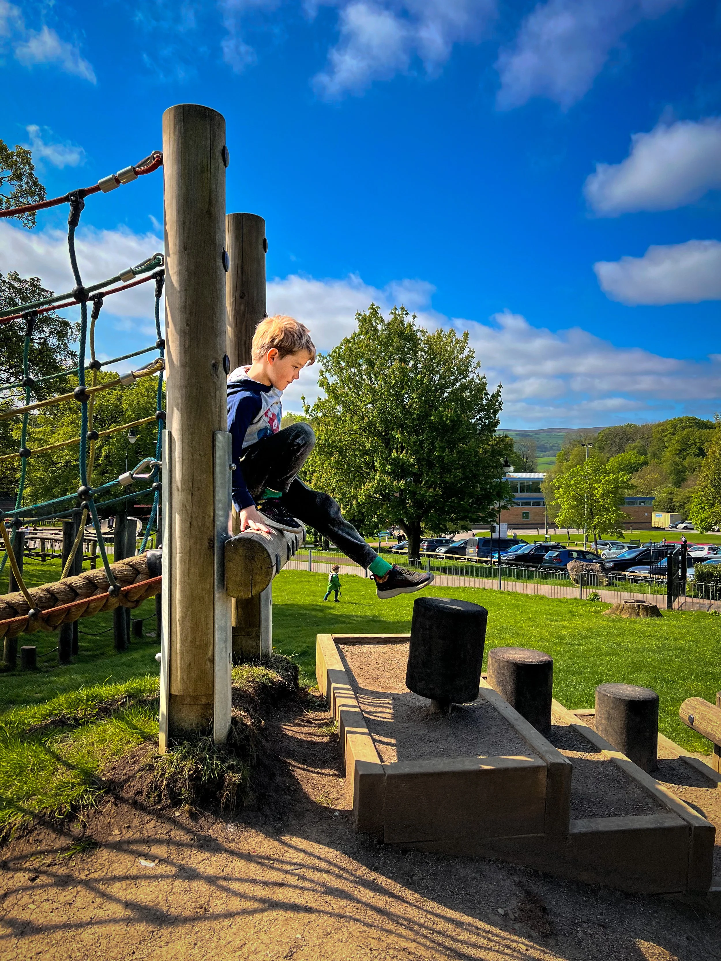 Child getting down from a rope bridge with a leisure centre behind with trees and hills