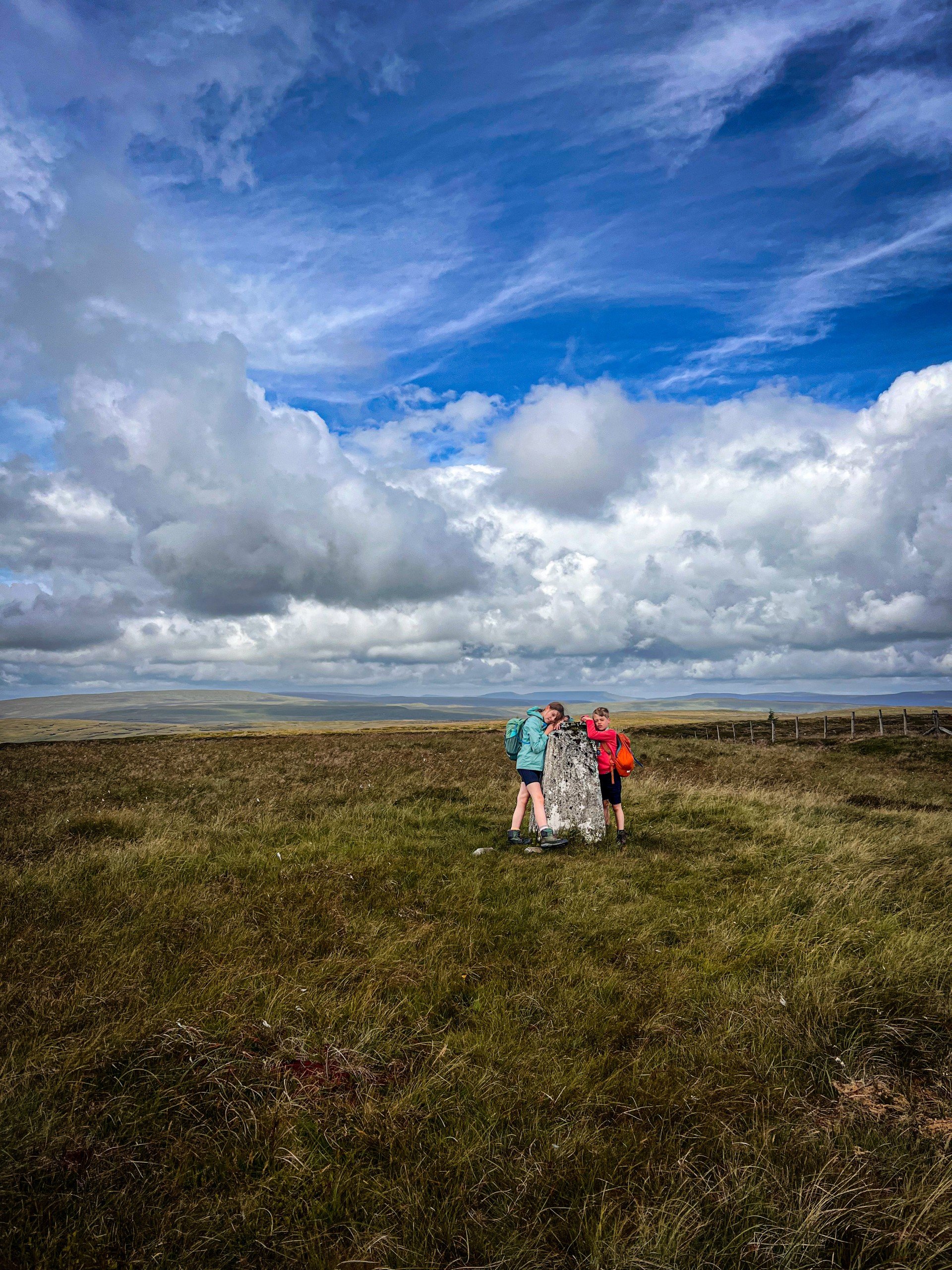 Children leaning on a trig point under blue skies on a Yorkshire Dales mountain