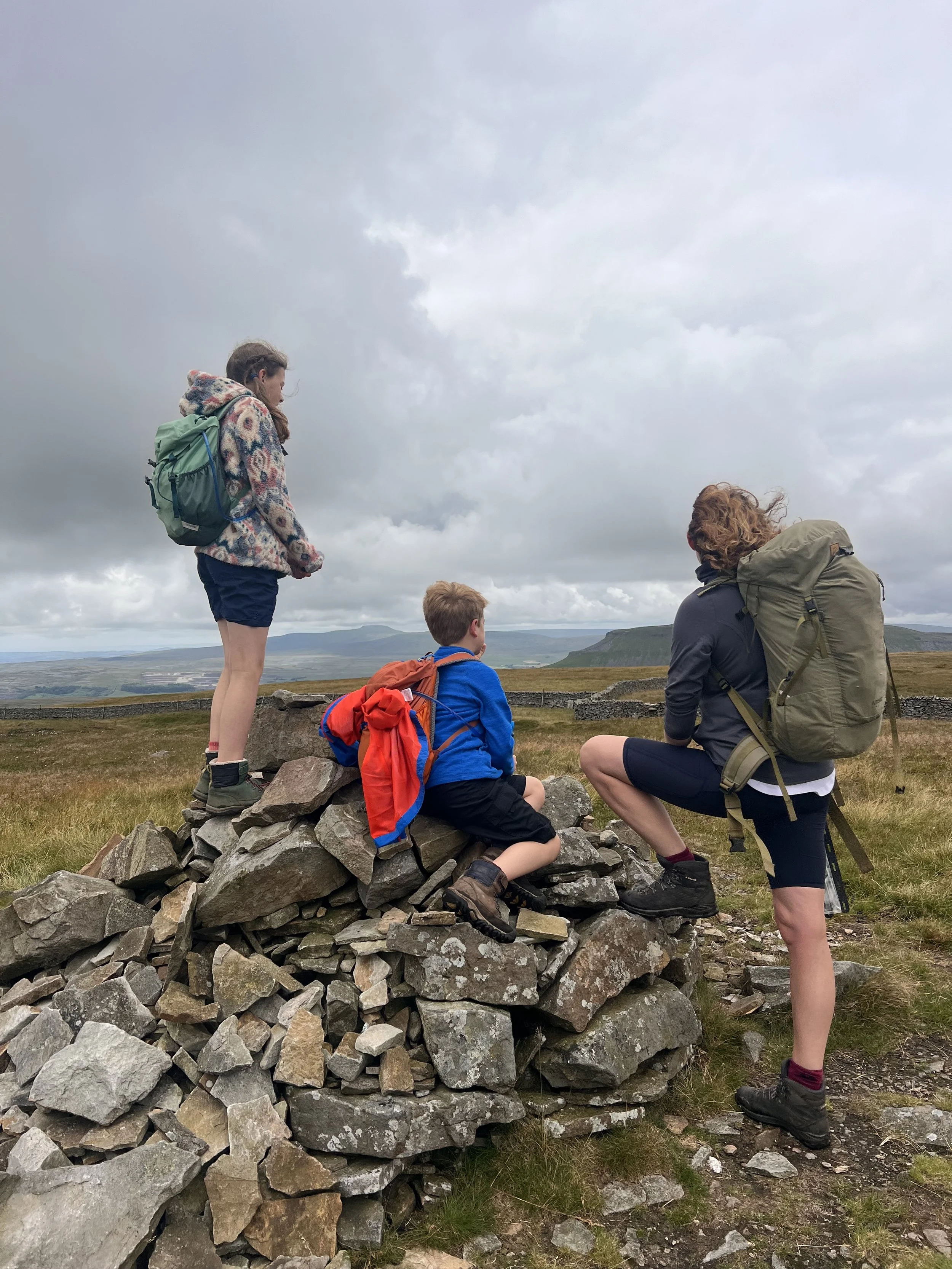 three people Sitting on a cairn with grey cloudy skies and mountains in the distance