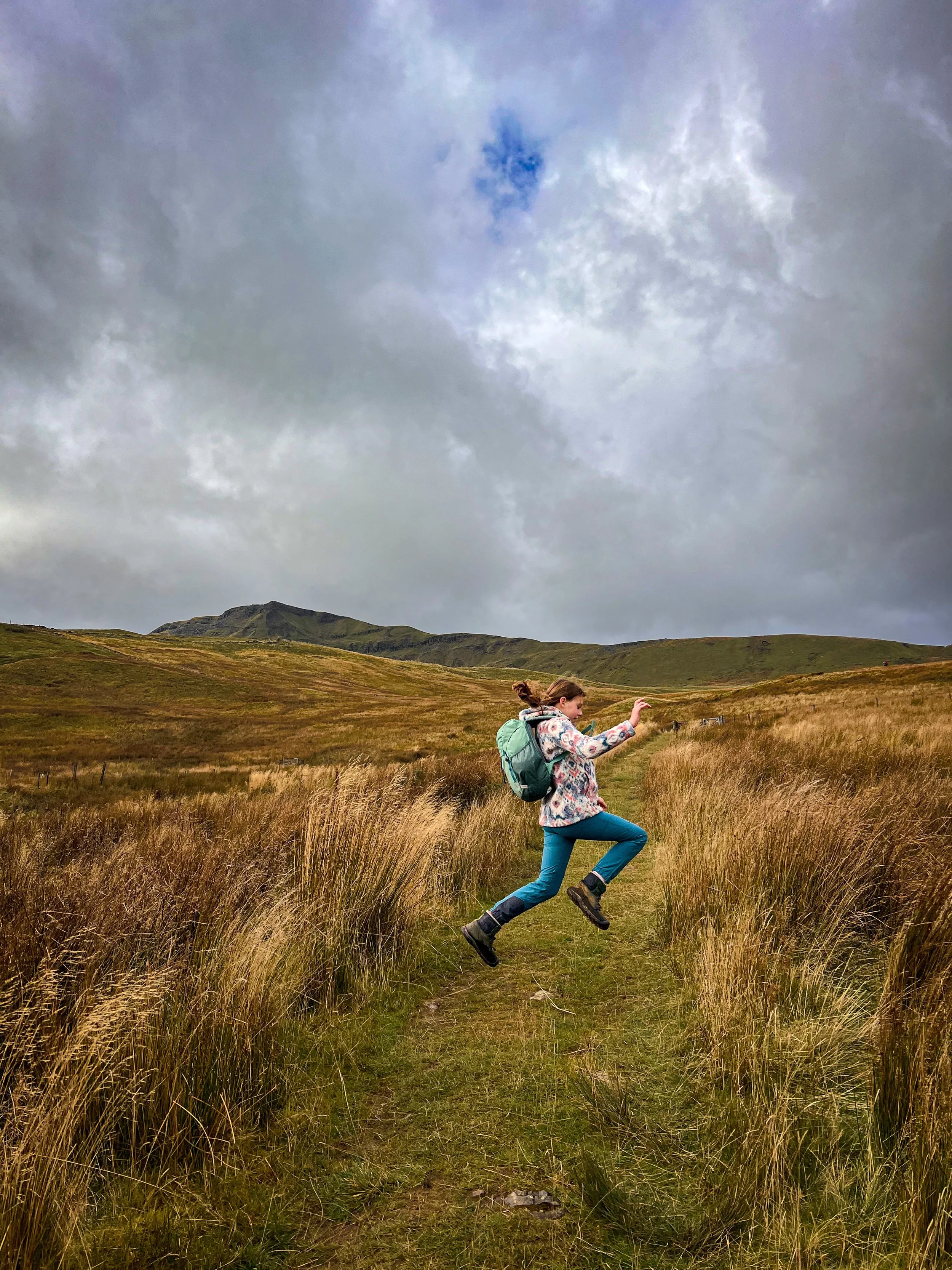 Child leaping on a path in the foreground with grass and with mountains behind