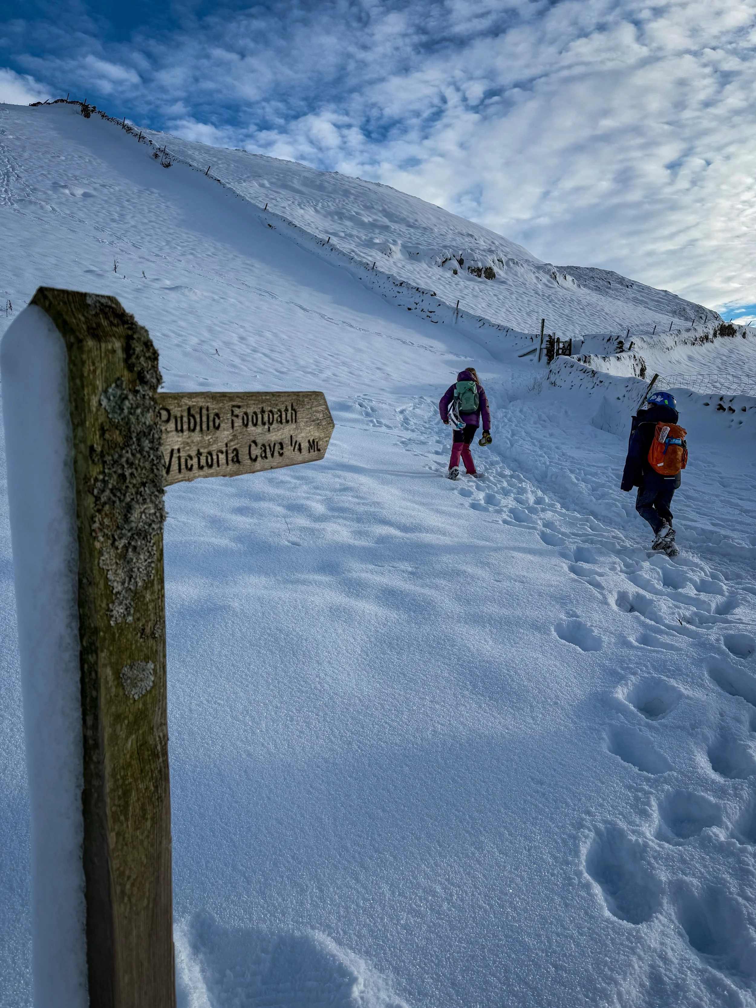 Children walking through snow with a public footpath sign in the foreground pointing to Victoria Cave