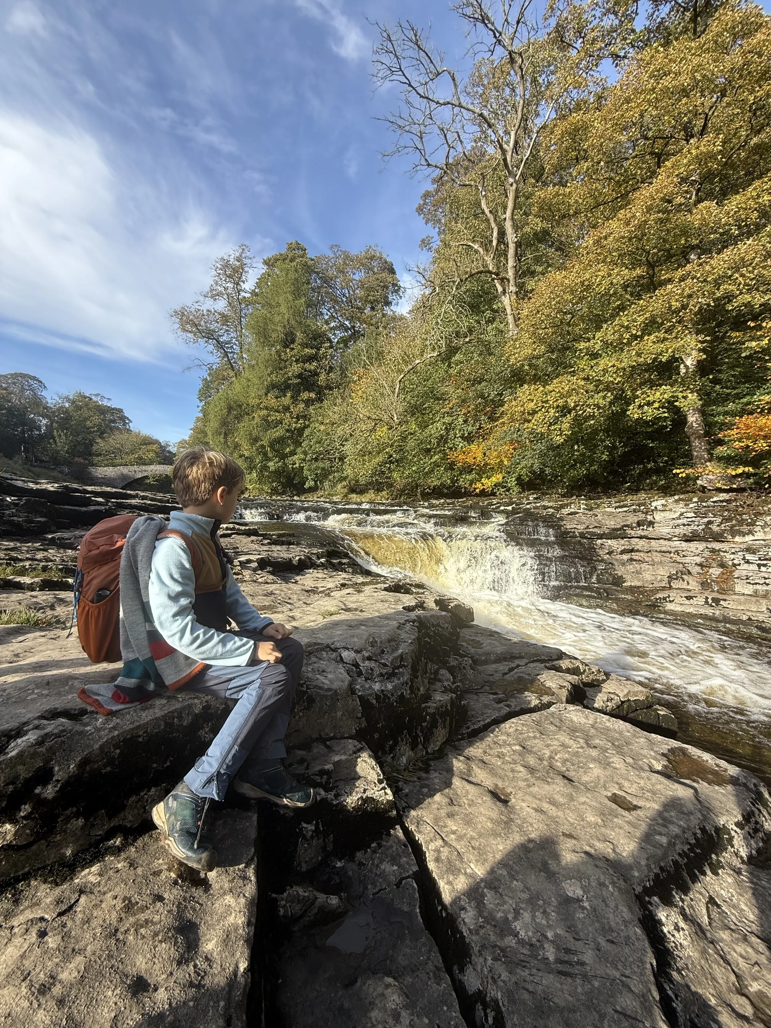 Child with early autumn trees sitting next to a waterfall under blue skies