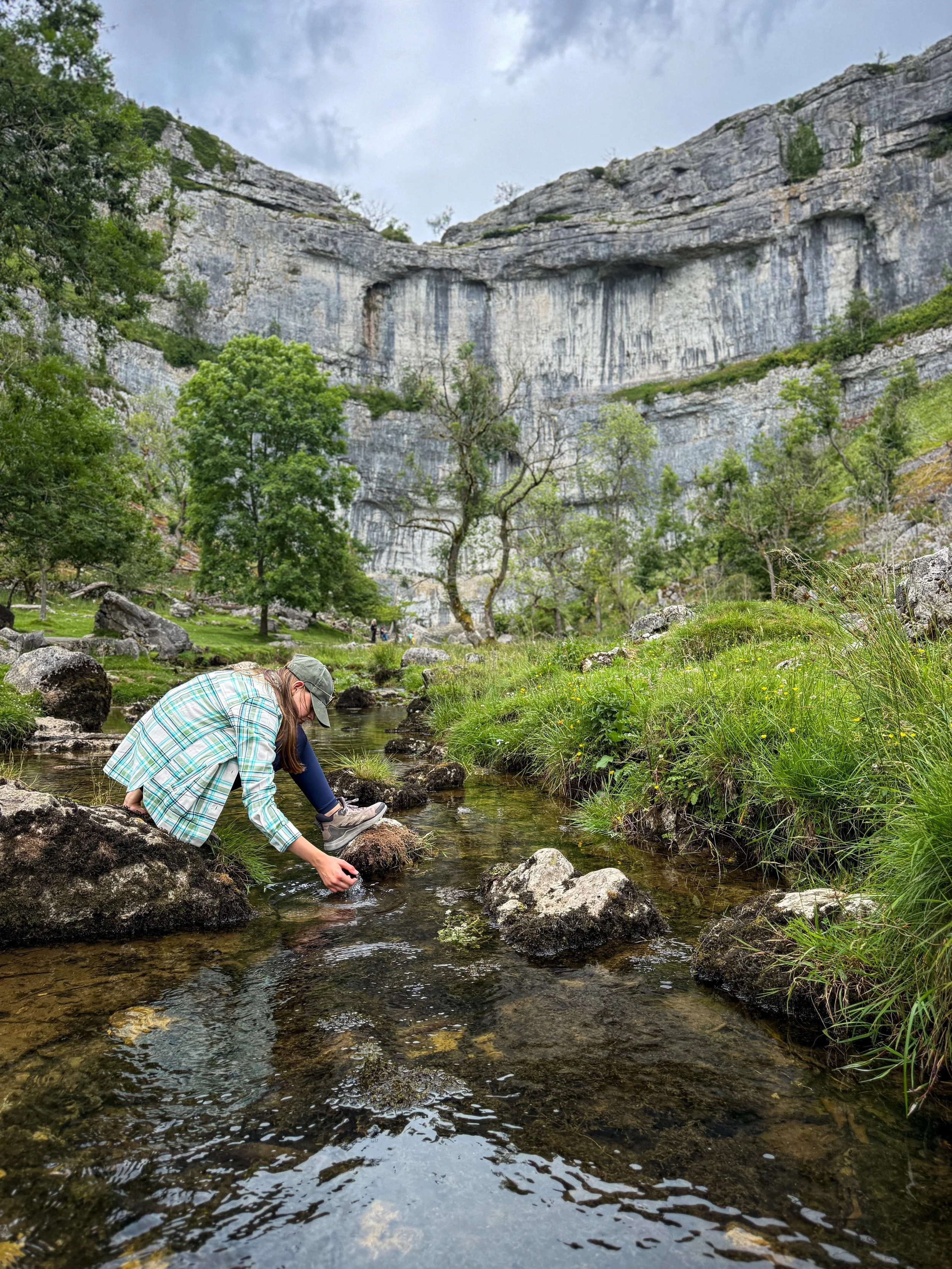 Child sitting on rock in the middle of a stream with hand in the water and malham cover cliff face behind