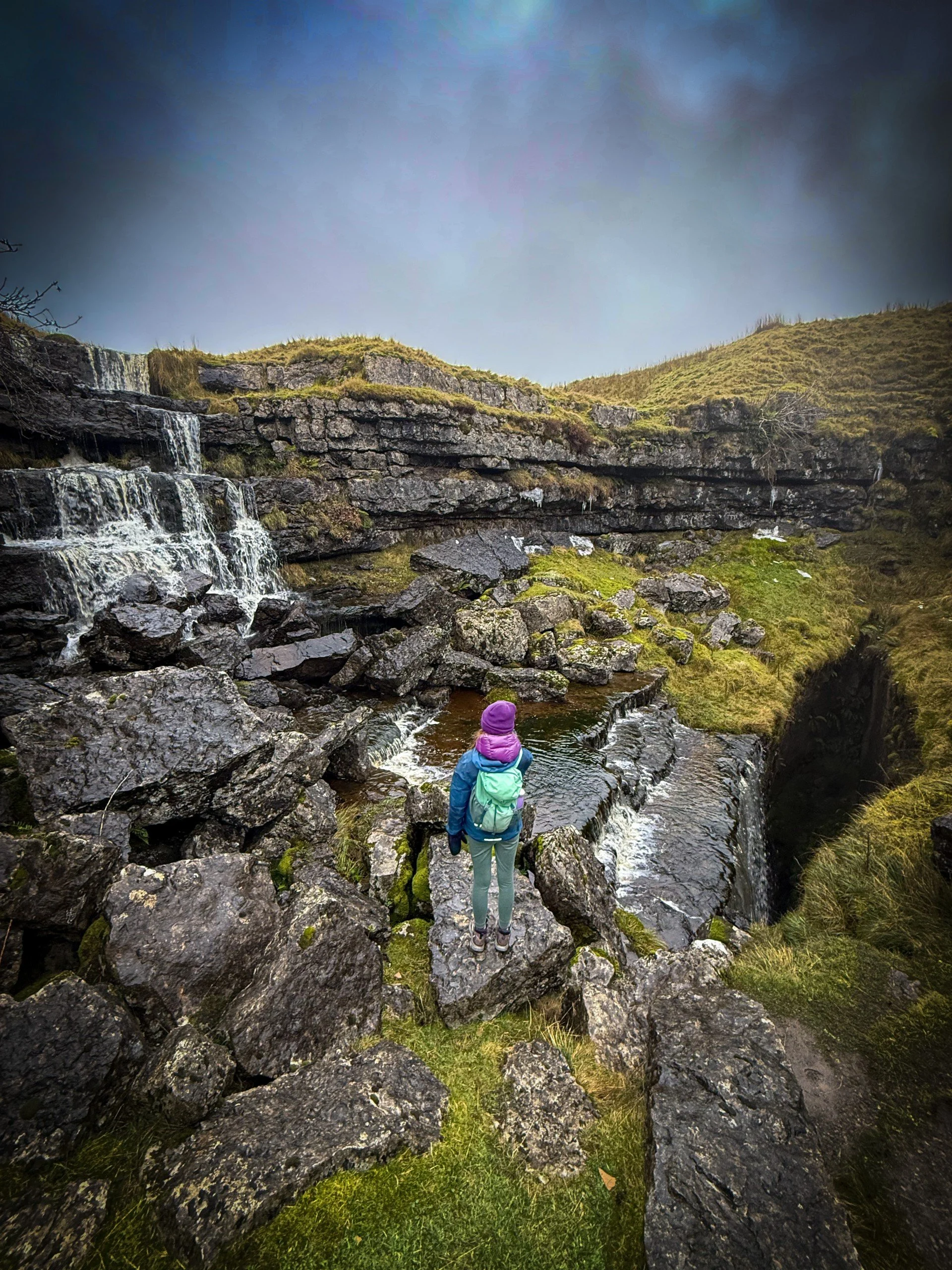 Child standing at the side of a waterfall with a large cleft in the ground on the right in winter with dramatic clouds