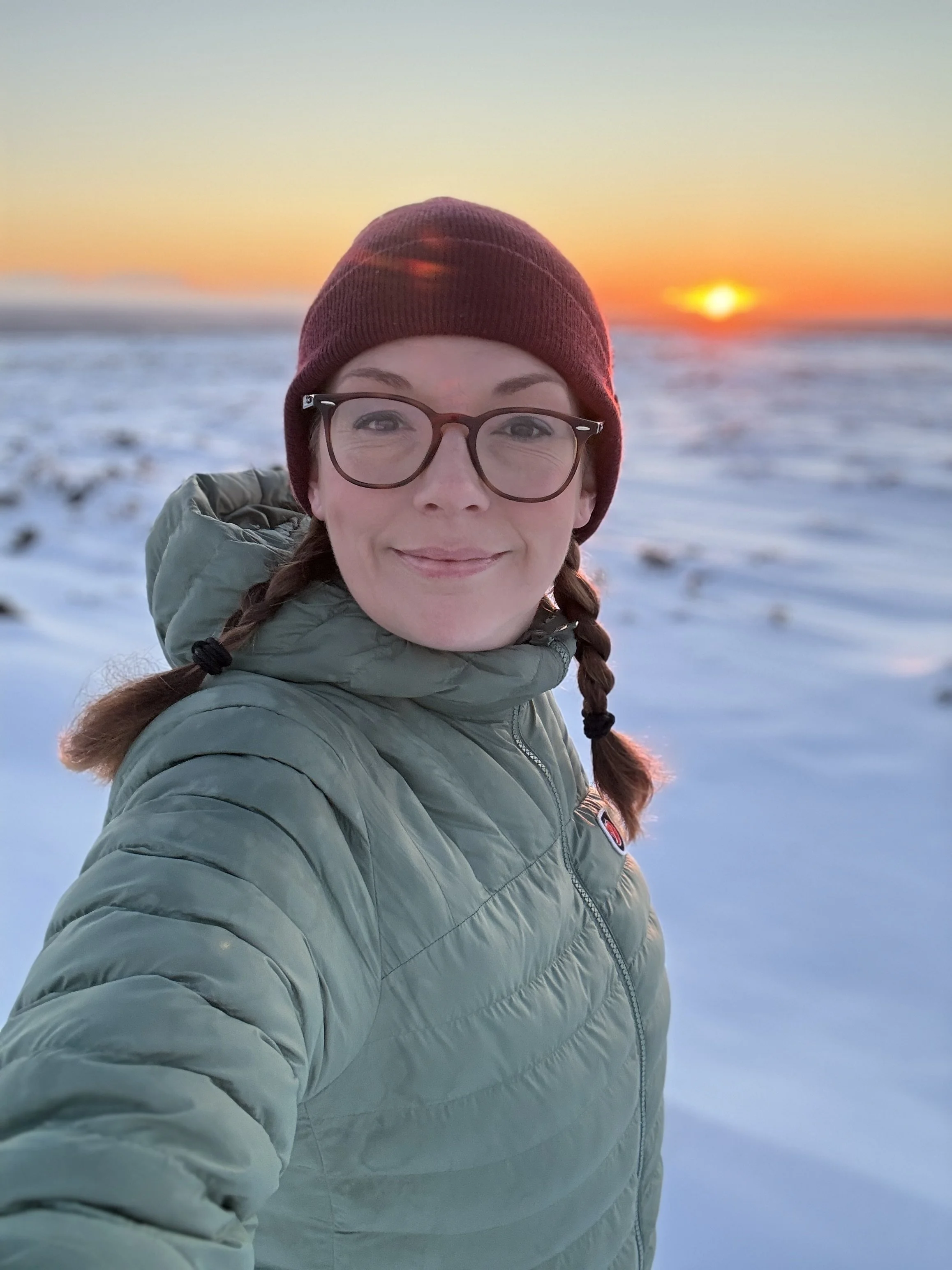 Mum with glasses on and a purple beanie wearing a sage green puffa coat in the snow on top of a moor with the sunrise behind