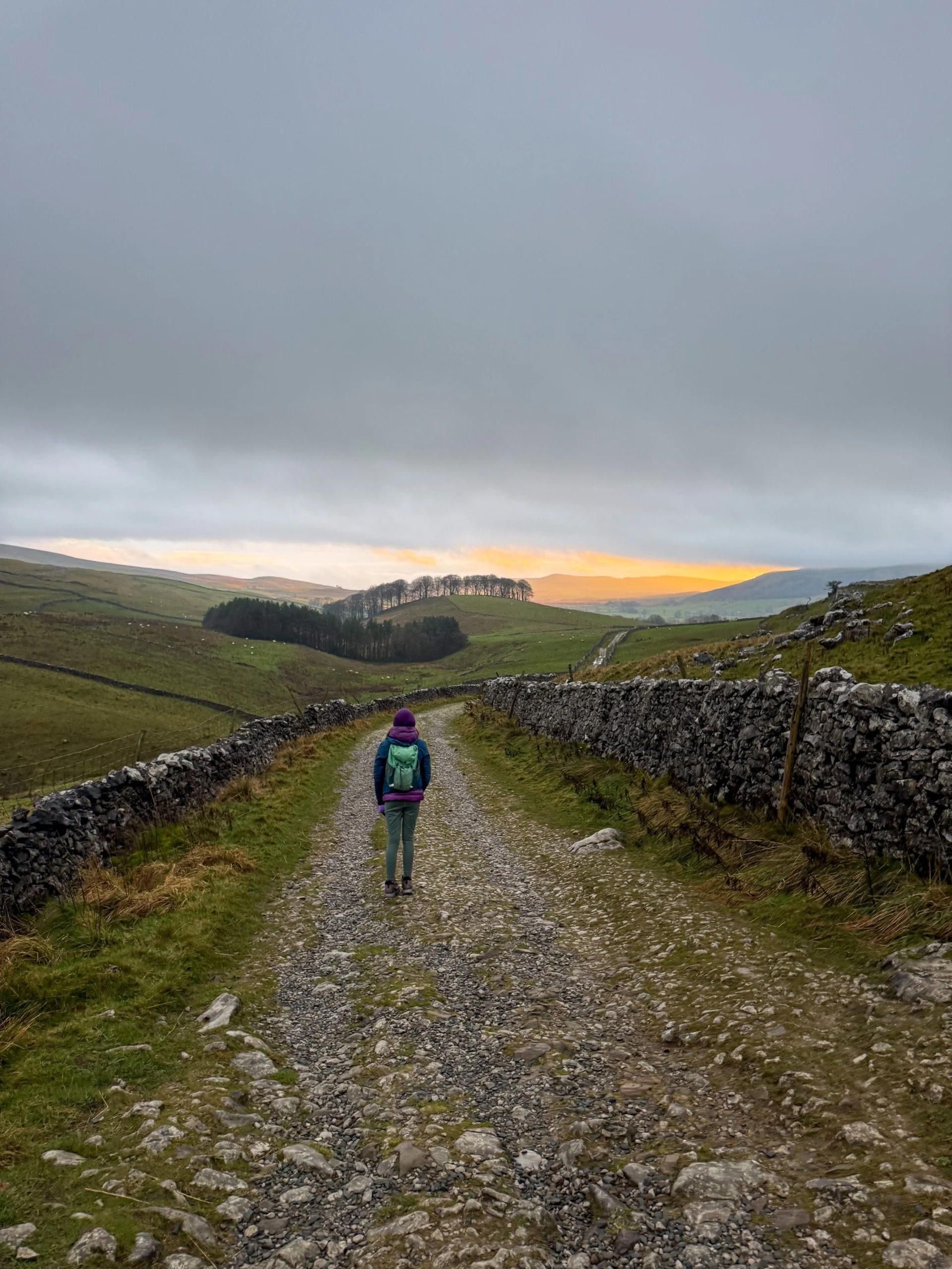 A glimpse of a winter sunset in grey clouds with a child walking down a dry stone walled track