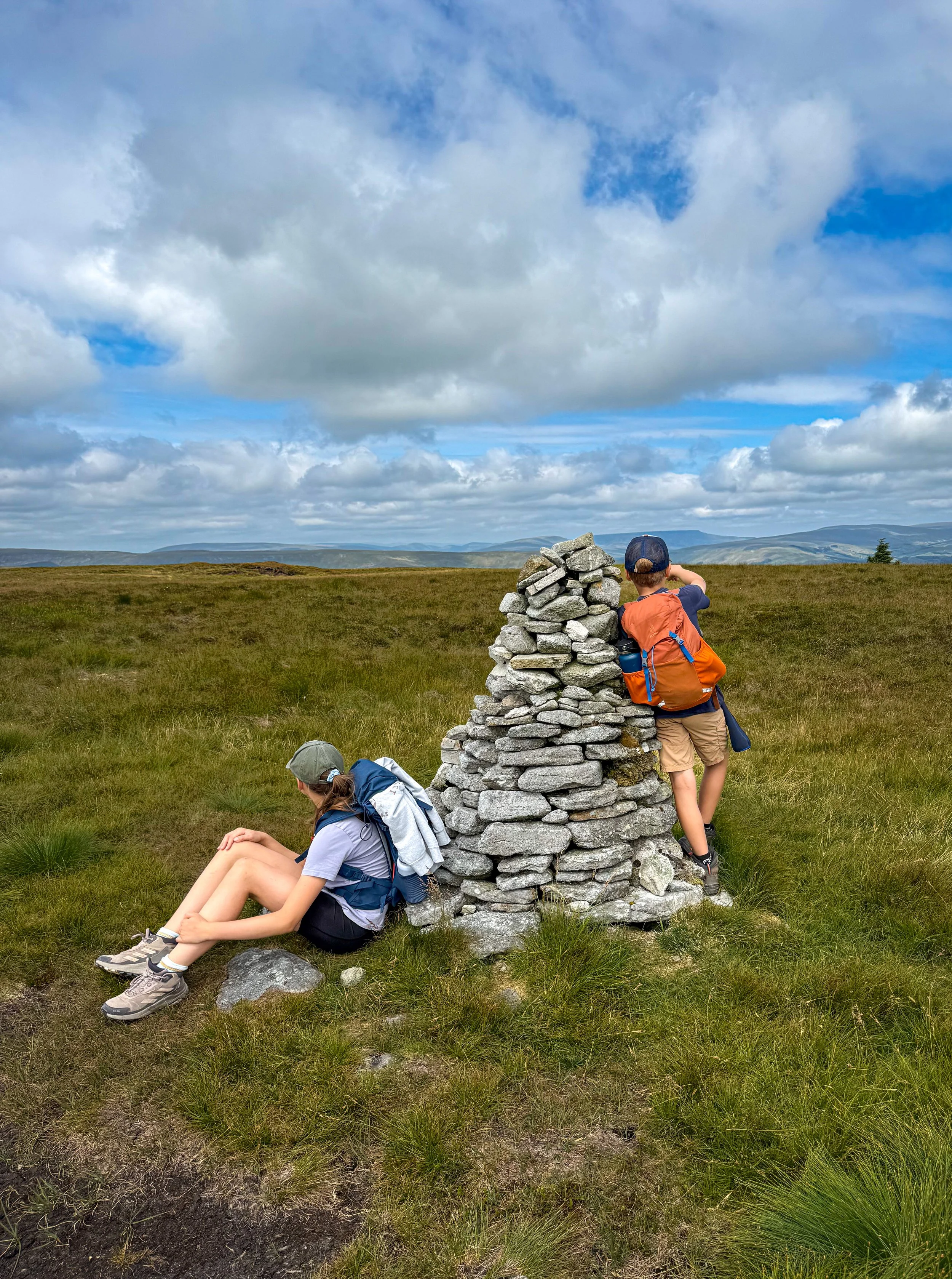 Two children wearing hiking gear next to a stone cairn one is pointing towards the mountains in the distance with blue sky and fluffy clouds above