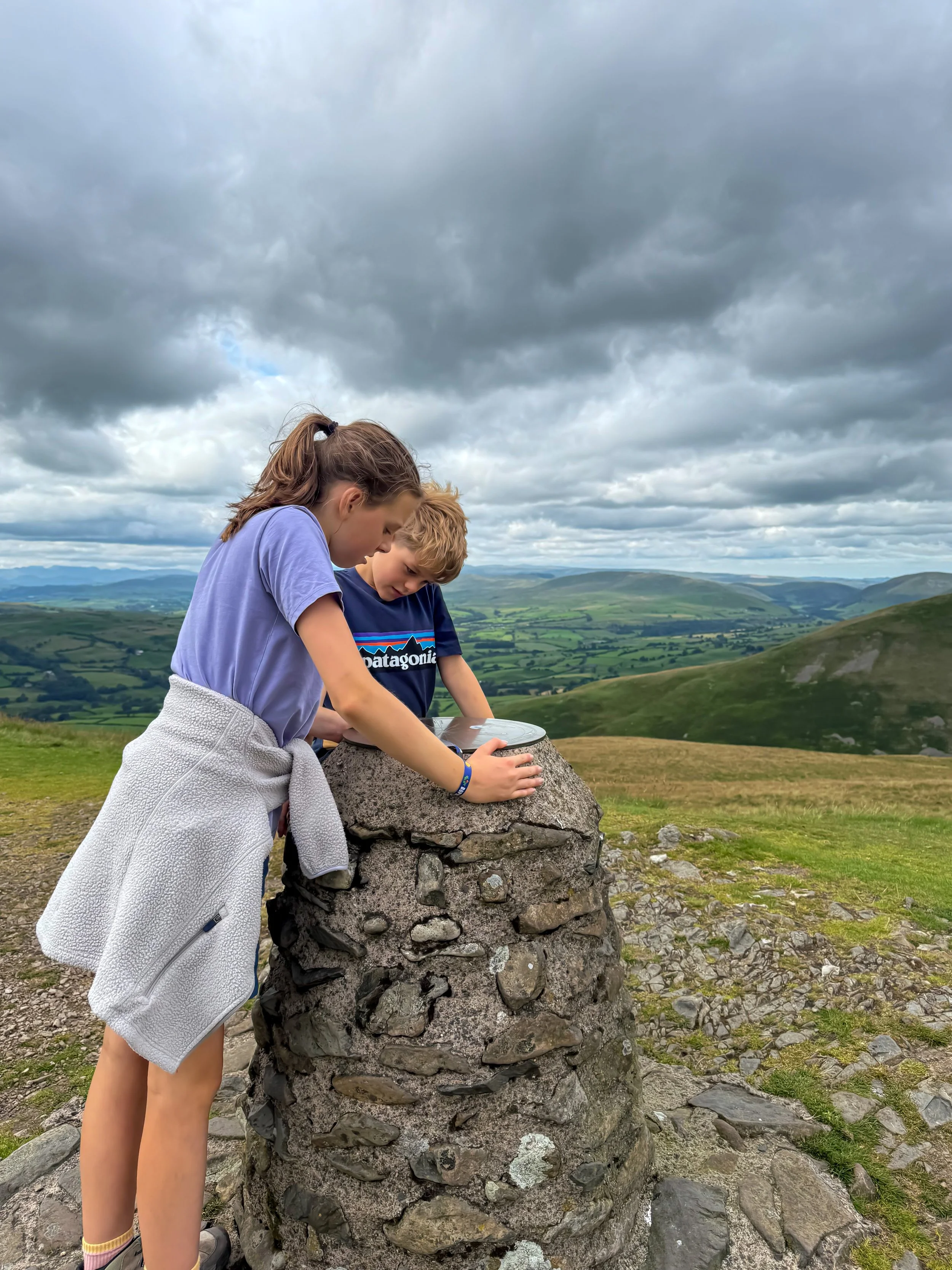 Children leaning over stone built pillar with a bronze plaque and hills behind