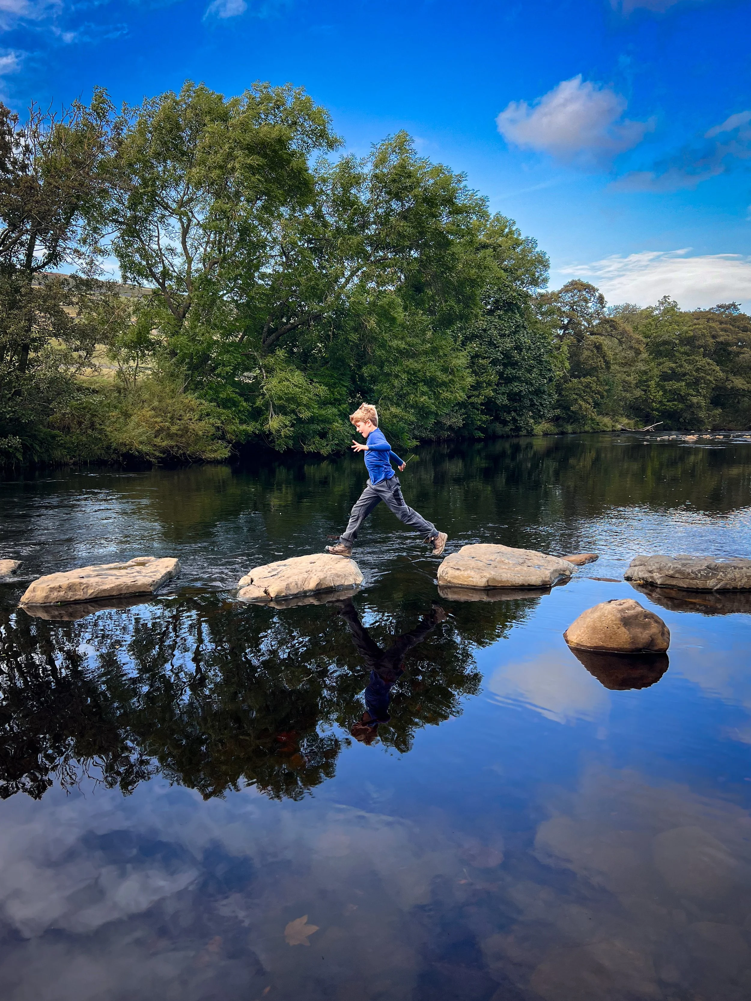 Child hopping over stepping stones across a calm river lined with trees in late Summer under a blue sky