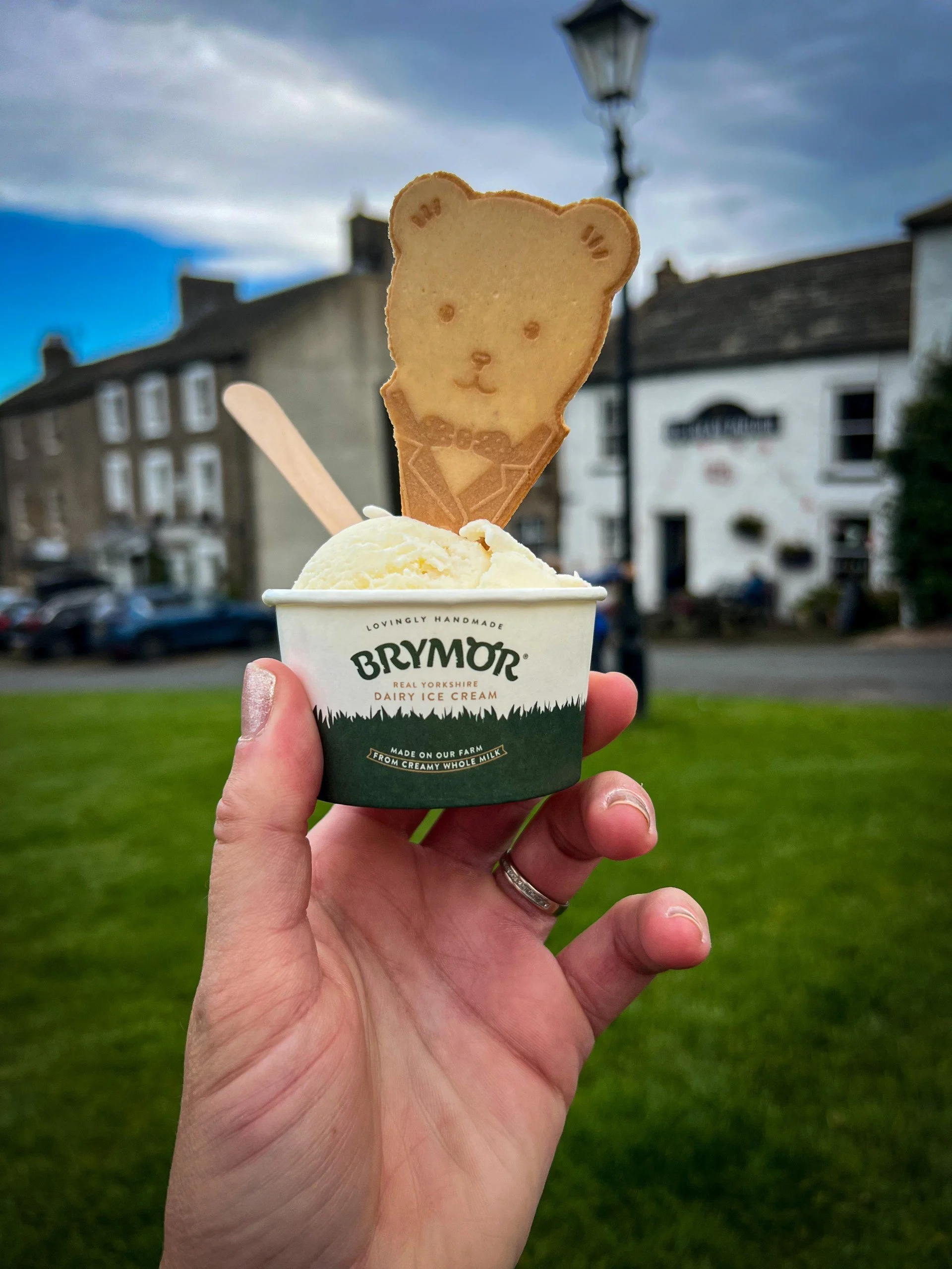 A hand holding up a small tub of Brymor ice cream with a teddy bear wafer in the top with  building behind a lamp post and blue skies