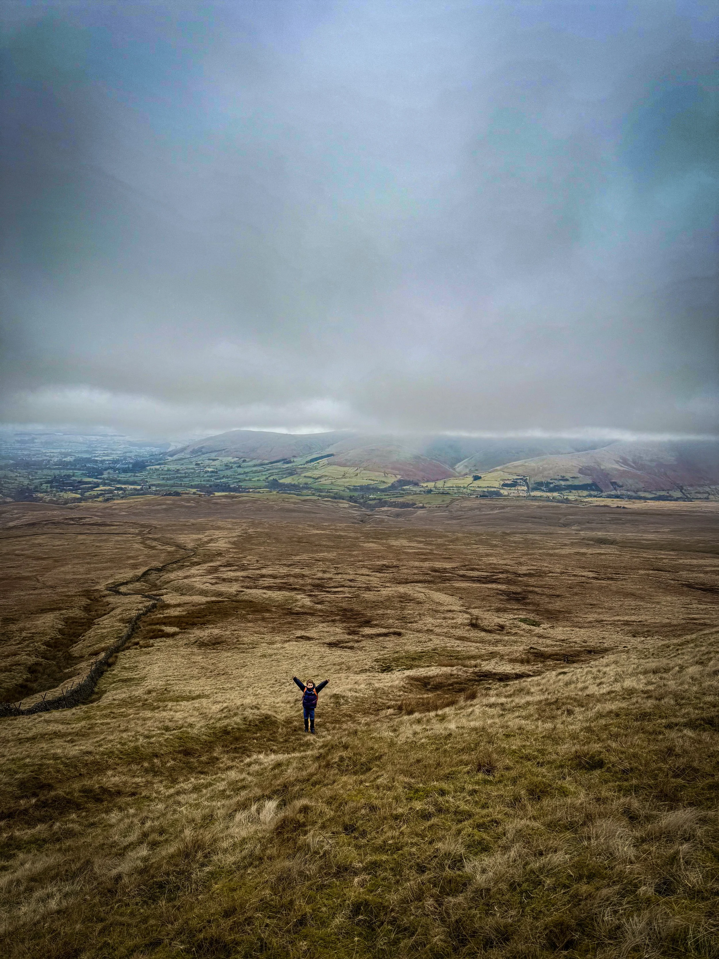 Child stood with arms in the air down a grassy hill with views of mountains beyond and low cloud