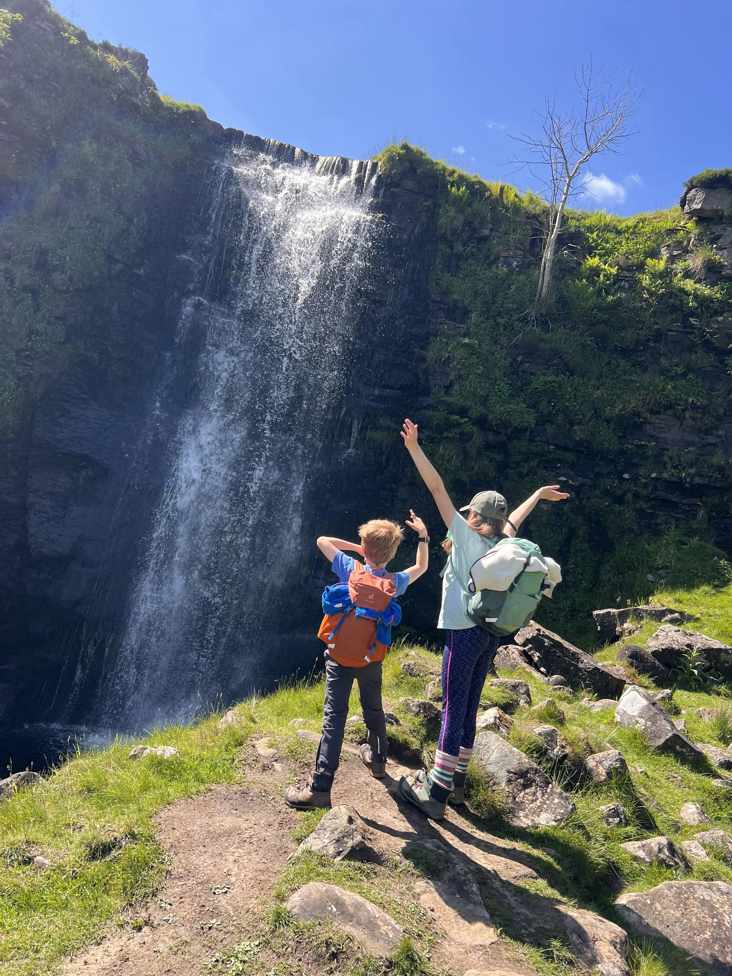Two children stood next to a waterfall with arms in the air in summer