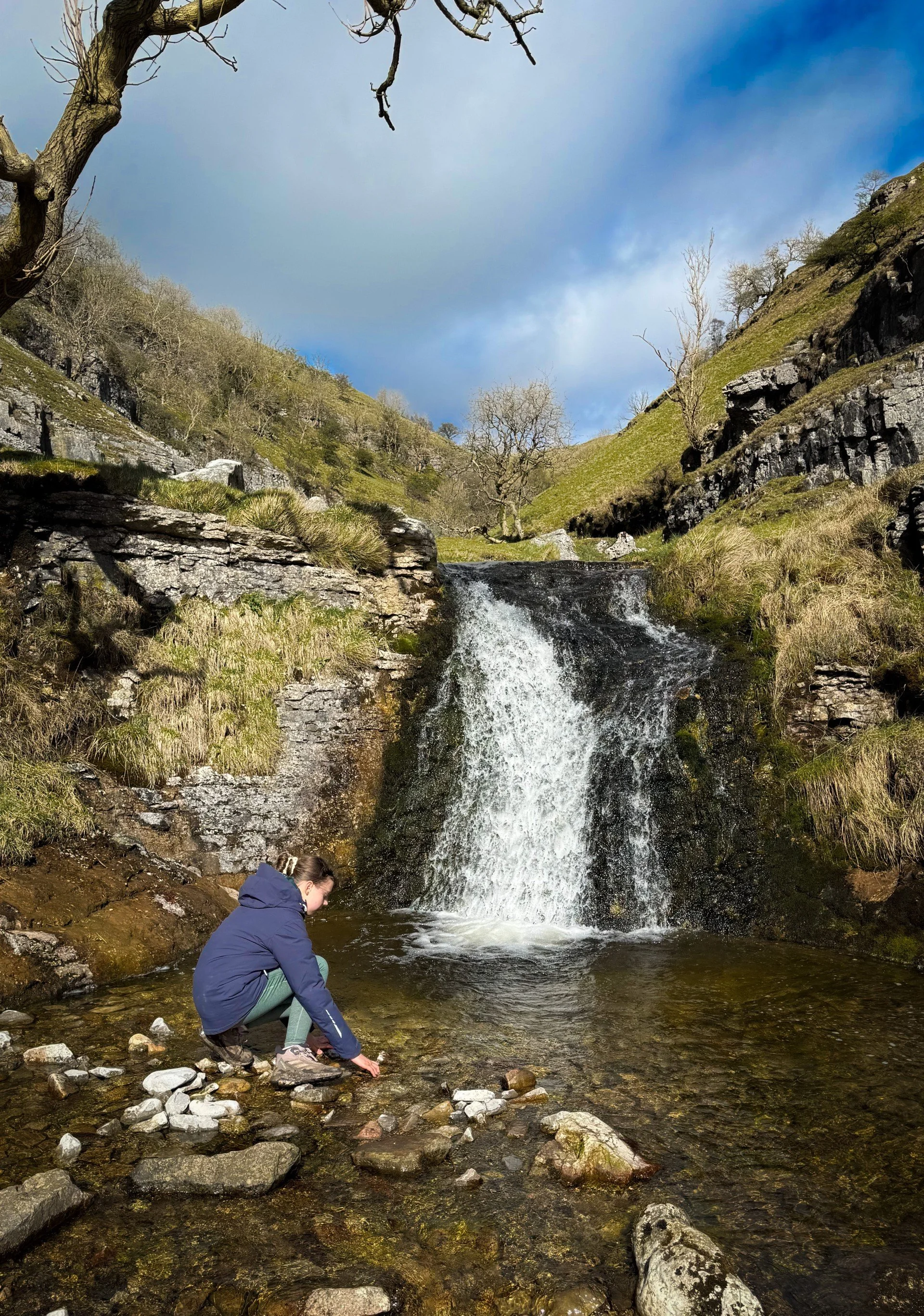 Child crouched in front of small waterfall playing in the water with clouds and blue skies above.