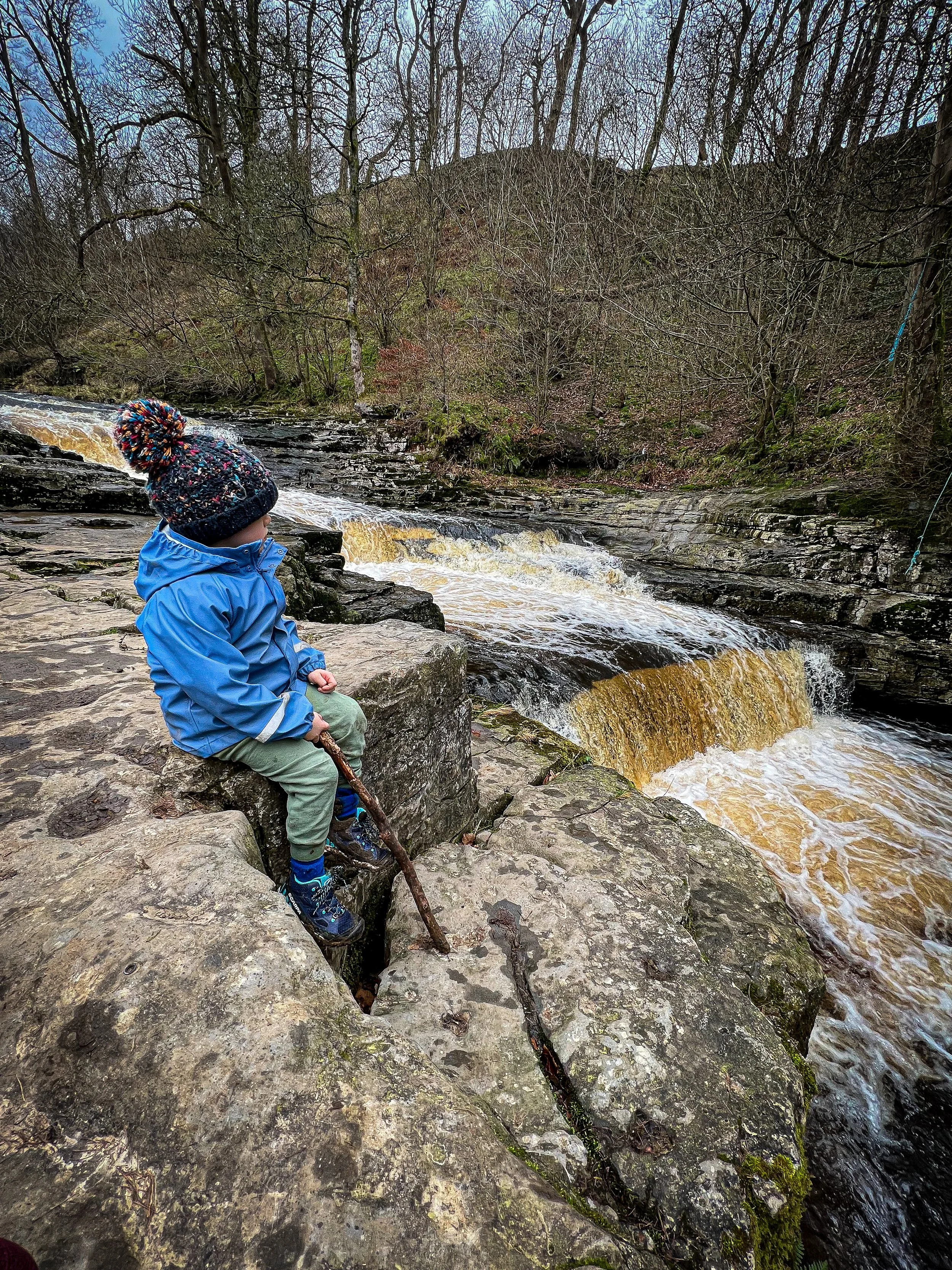 Child sat next to steppes waterfall with a stick wearing a blue coat and hat in winter