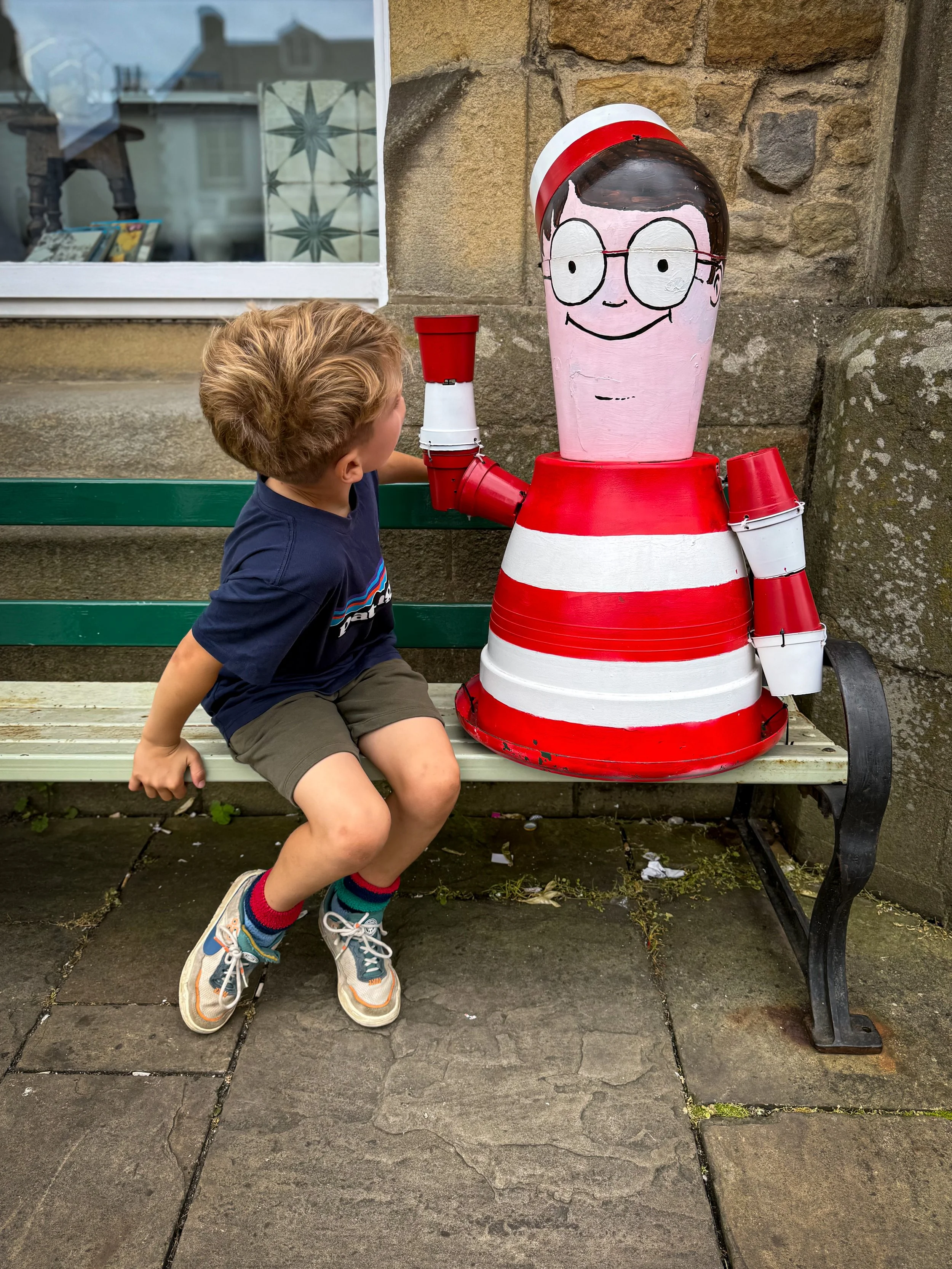 Child sat on a bench looking at a where's wally made of flowerpots