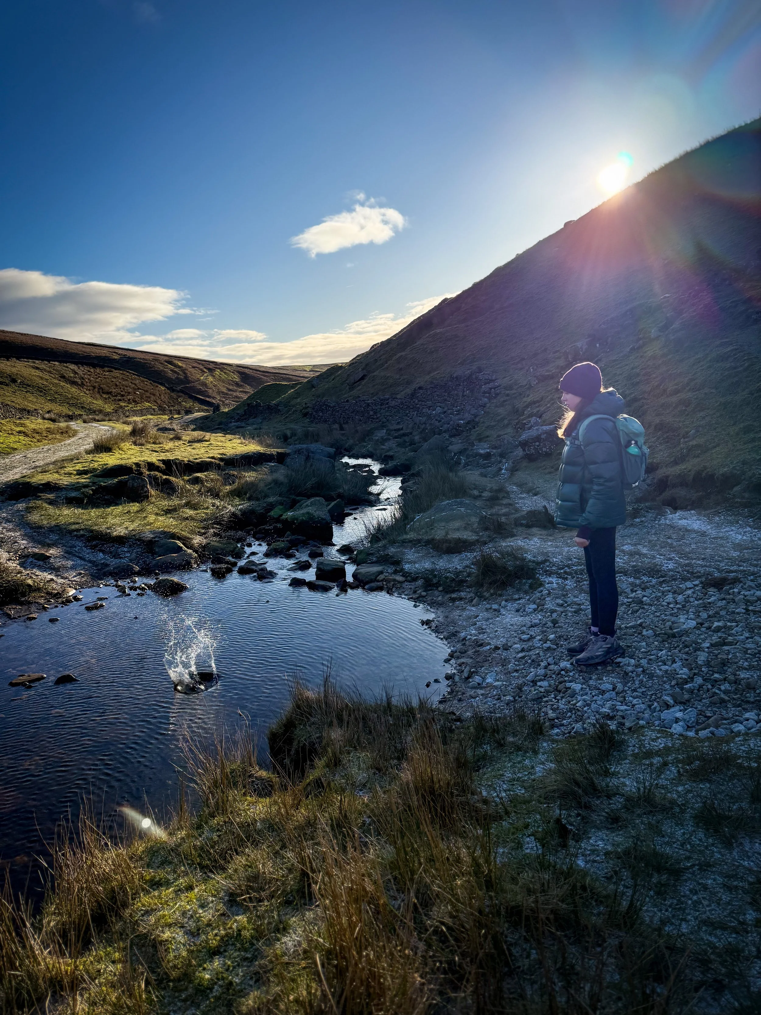 Child throwing a stone into the beck with blue skies and sun behind the splash of water is visible.