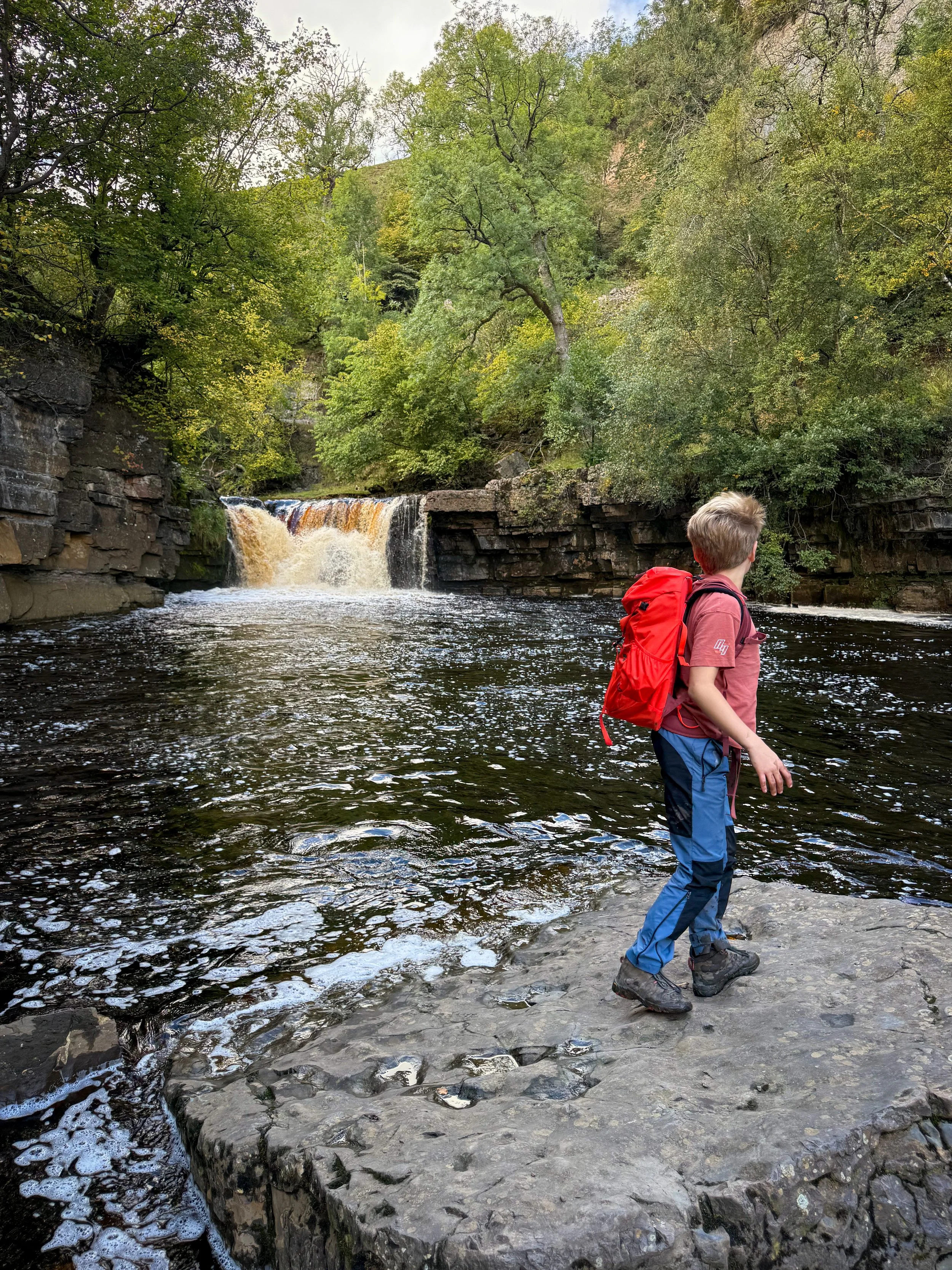 A child standing on a large rock in front of a waterfall on a river which is very full with autumn trees around
