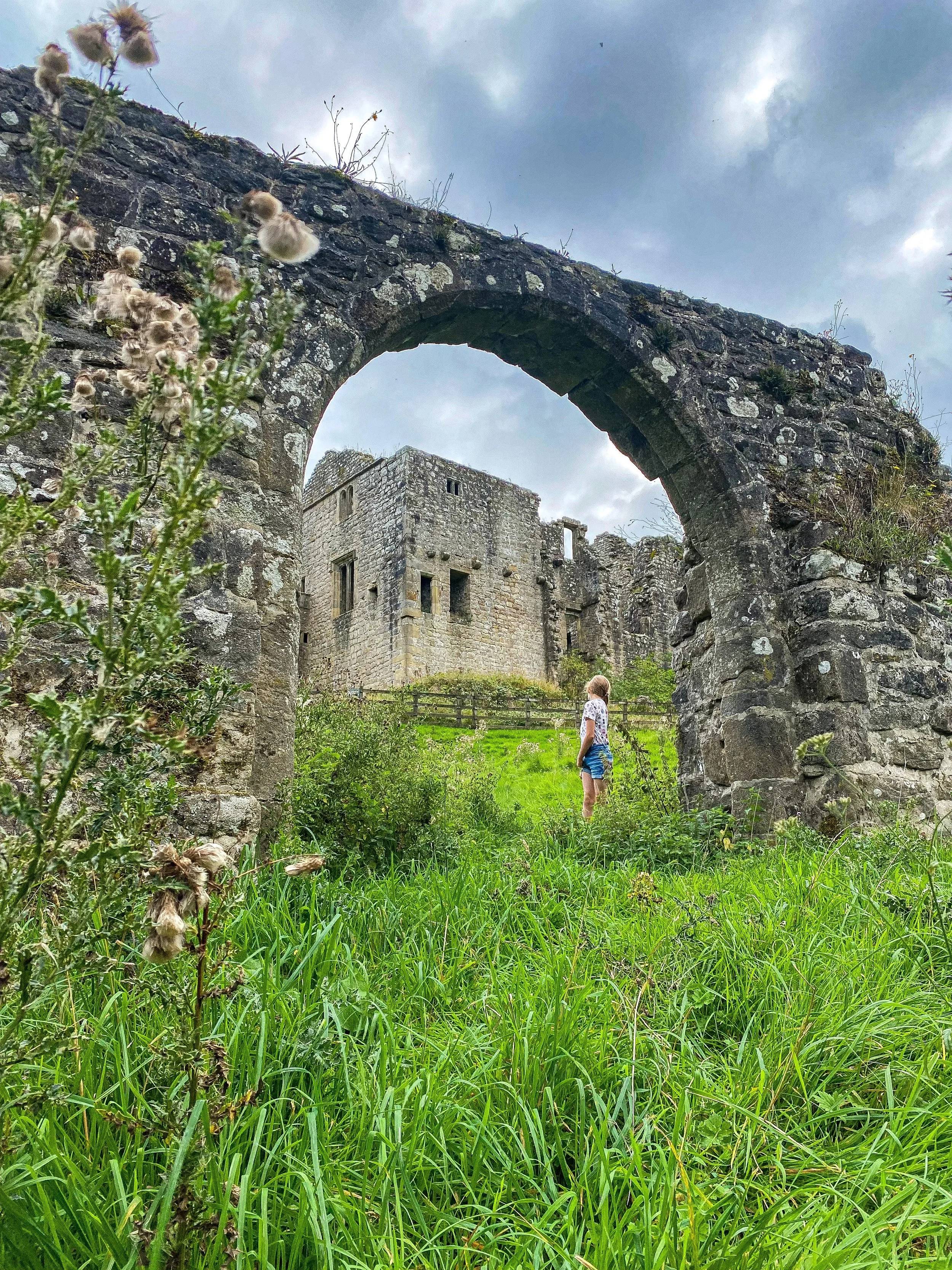 Child in a stone archway with ruins of an old hunting lodge behind with green grass and late summer thistles