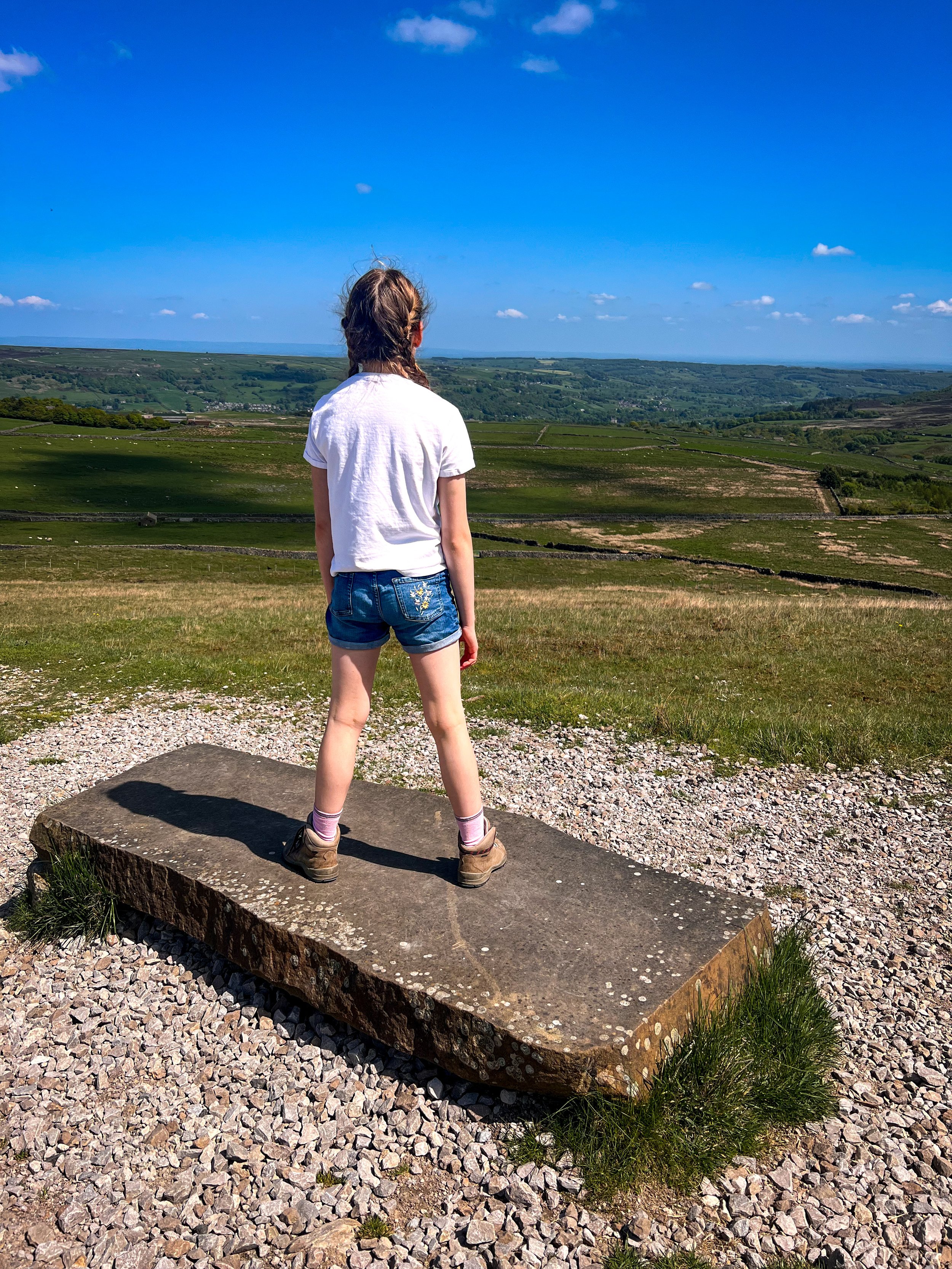 Child standing on a rock under blue skies in Summer looking at view of Nidderdale