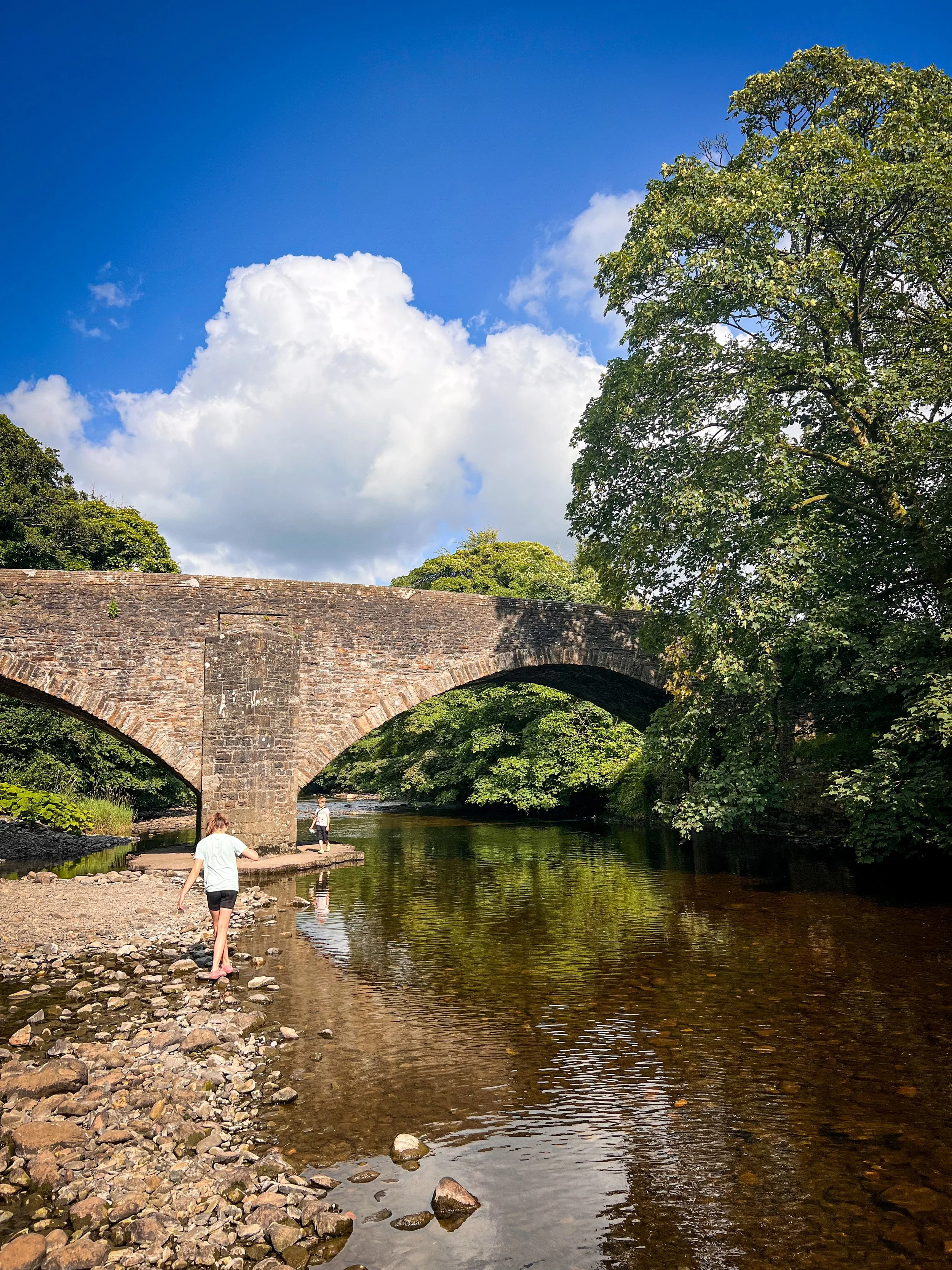 Child walking along the edge of a river with a bridge behind under blue sky