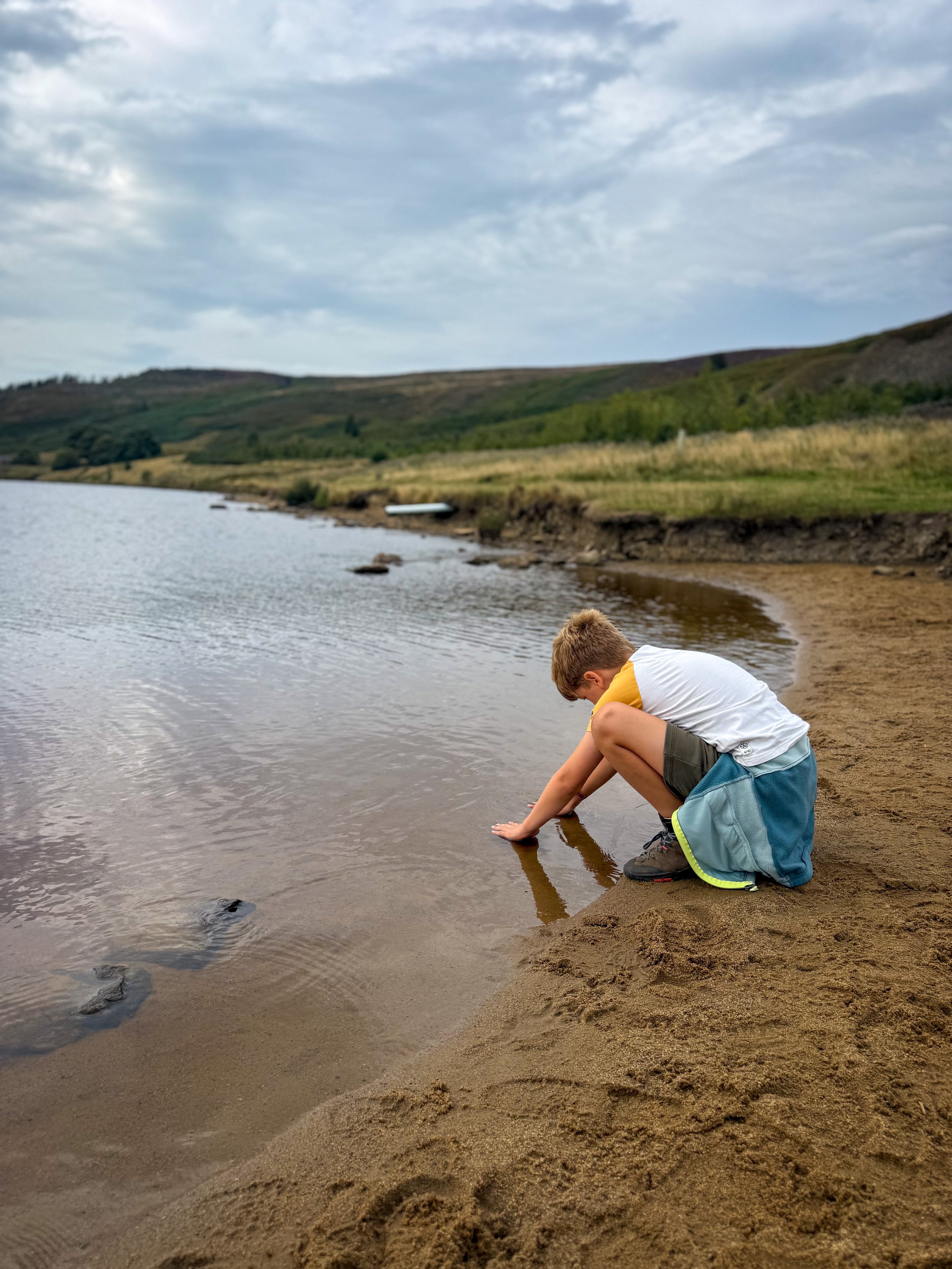 Child with hands in the water of a reservoir with grassy banks beyond