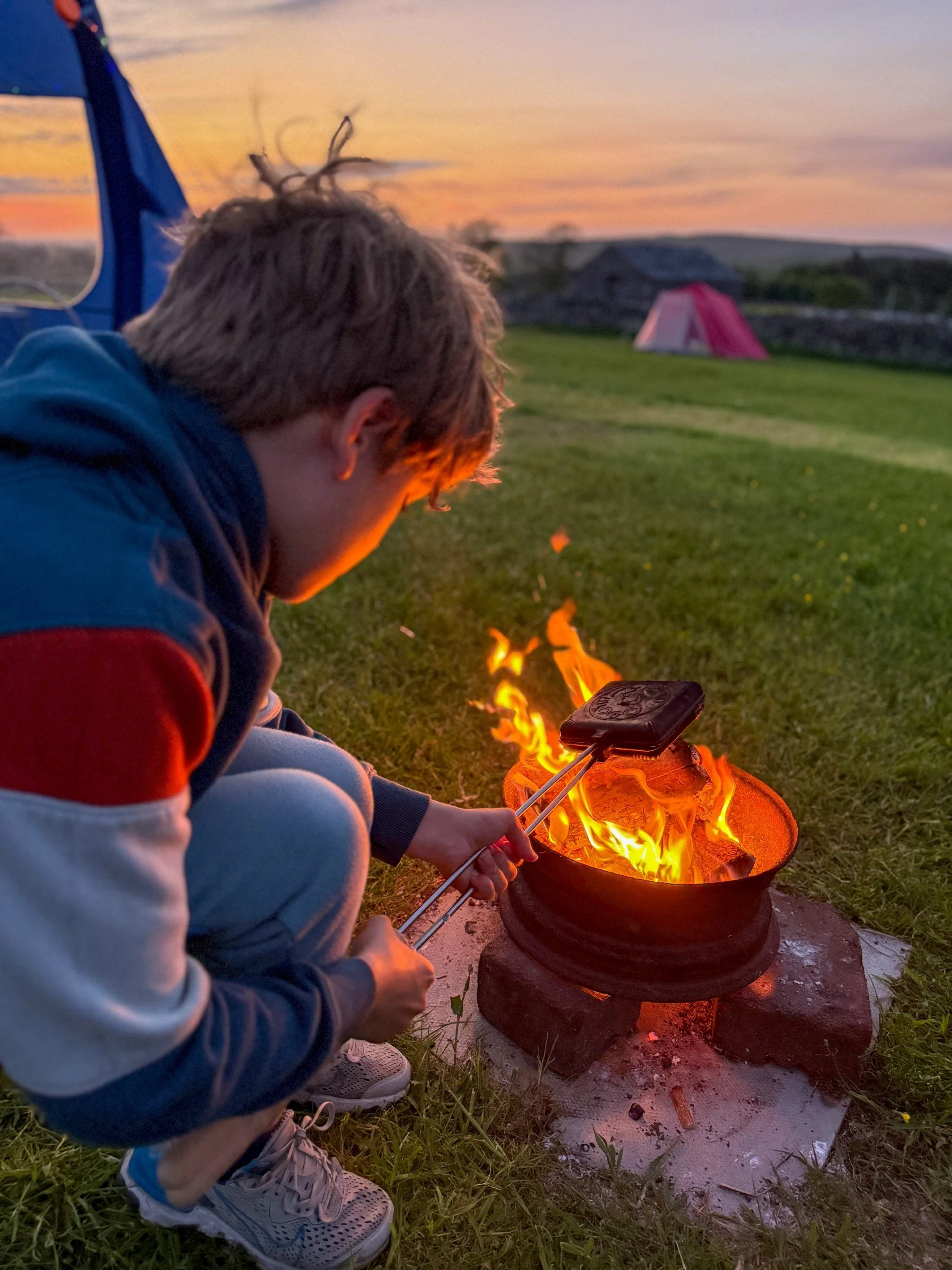 Child sitting next to campfire with tent and sunset behind