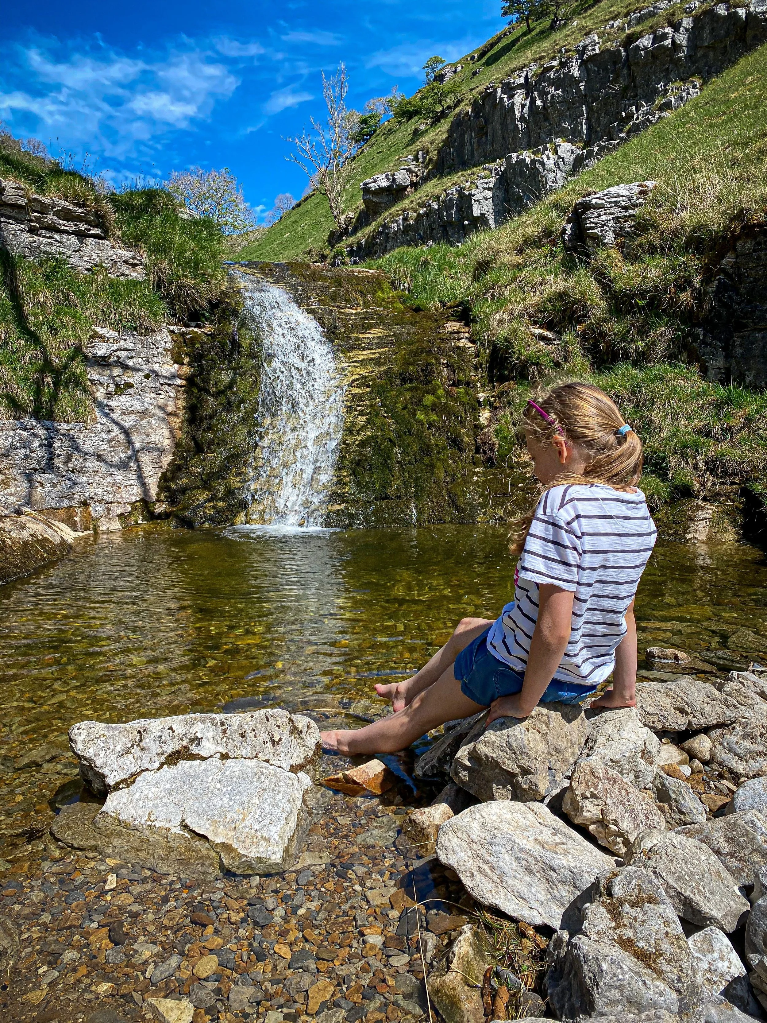 Child sat perched on rock with toes in the water next to a waterfall cascade with grassy and rocky slopes beyond under blue skies