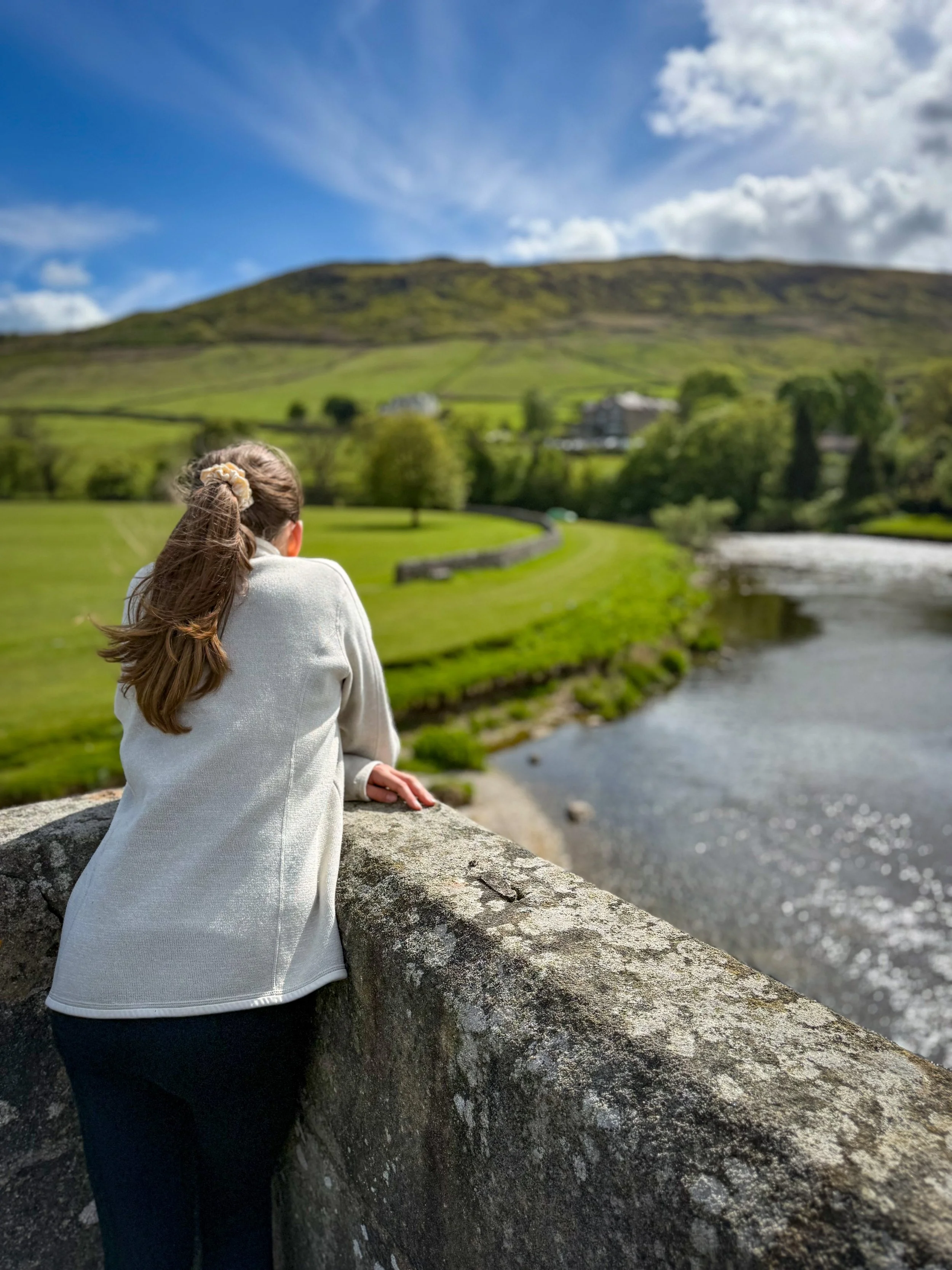 Child leaning over an old stone bridge looking down at the river with fells behind