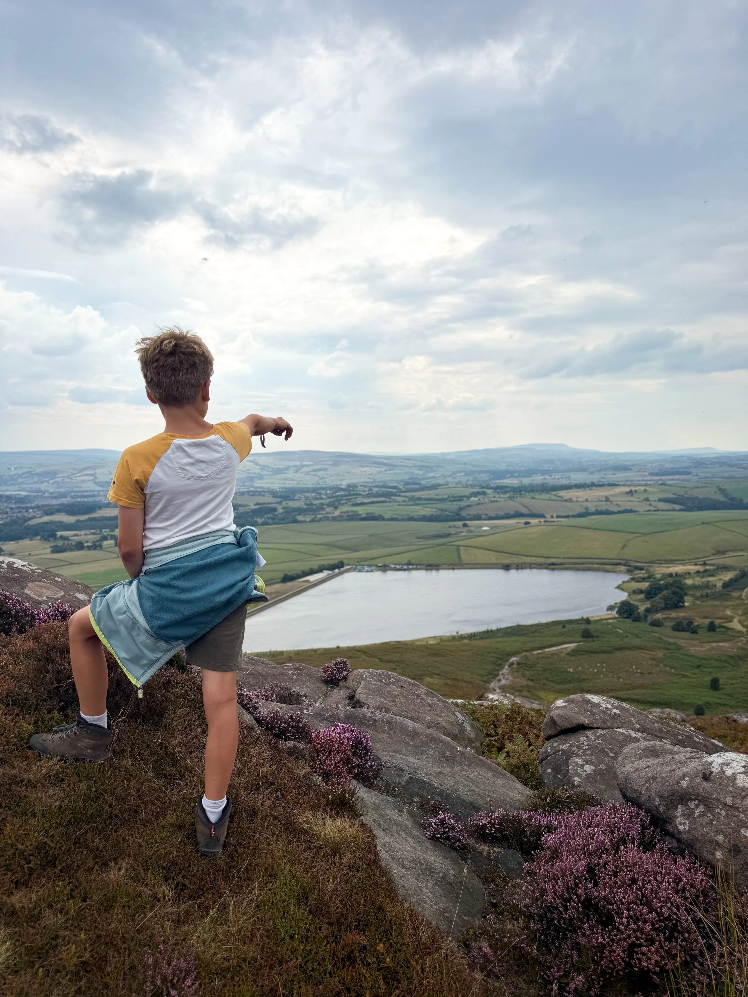 Child standing on a rocky summit with purple heather pointing out towards a reservoir with hills in the distance