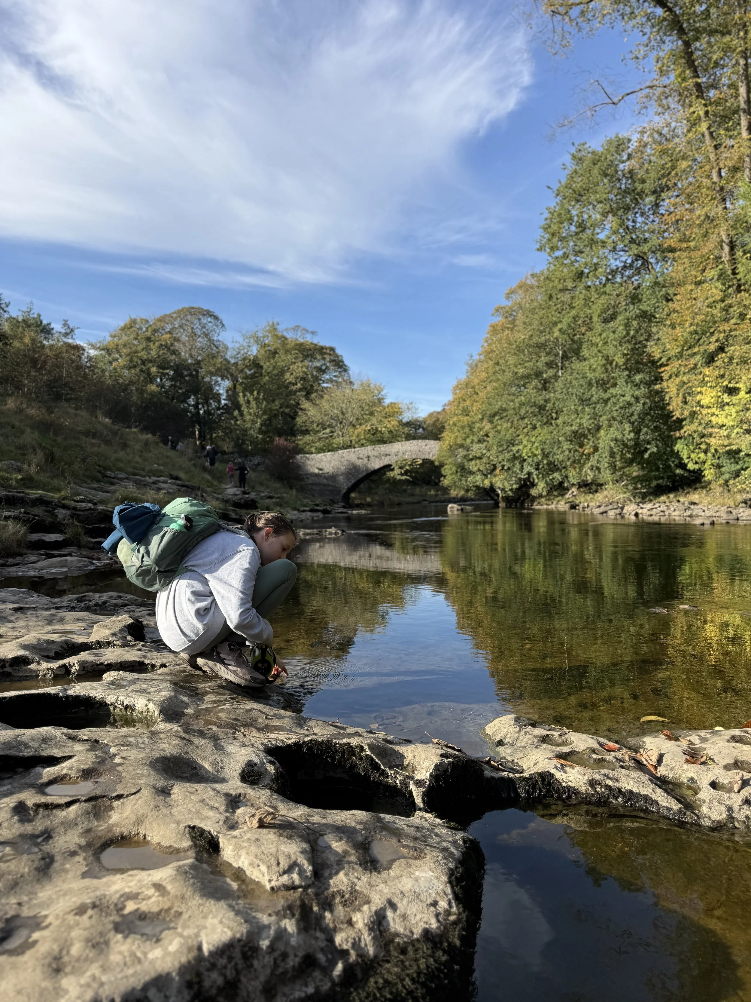 Child playing in calm water with a bridge behind and autumn leaves on the trees with blue sky