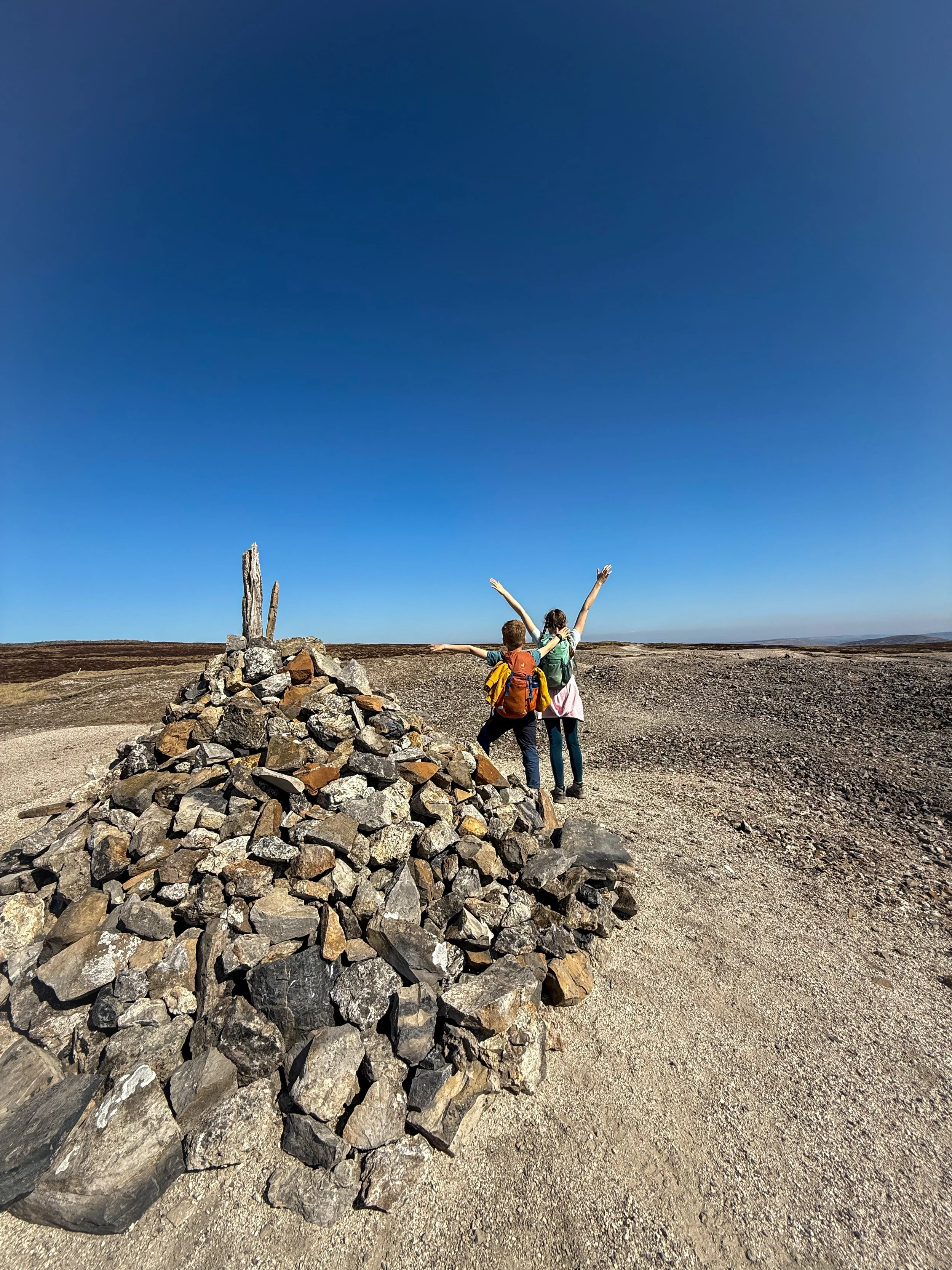 Children with arms in the air standing next to large cairn with spoil heaps and blue skies