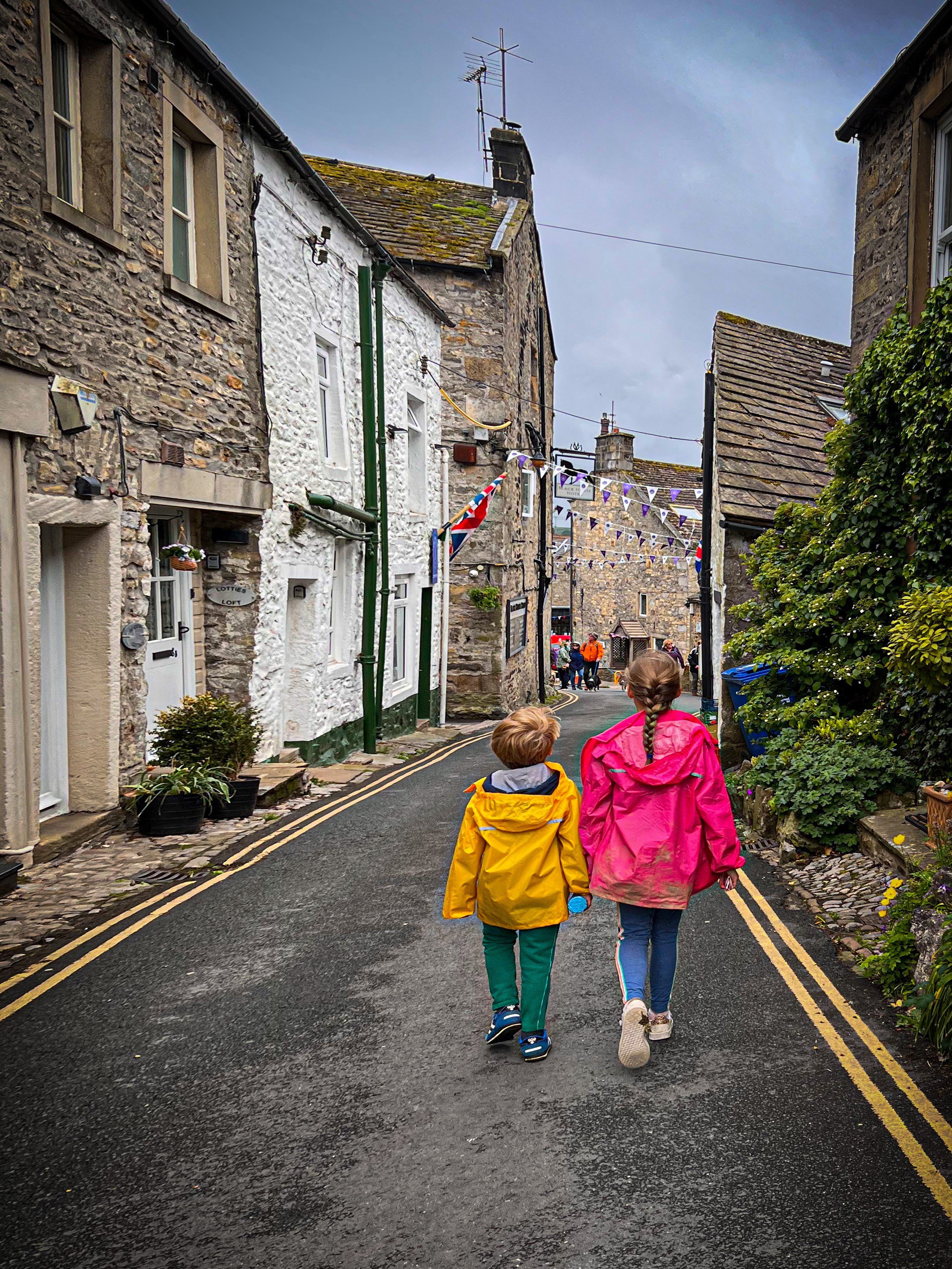 Two children in pink and yellow raincoats walking down a traditional narrow country street in Grassington